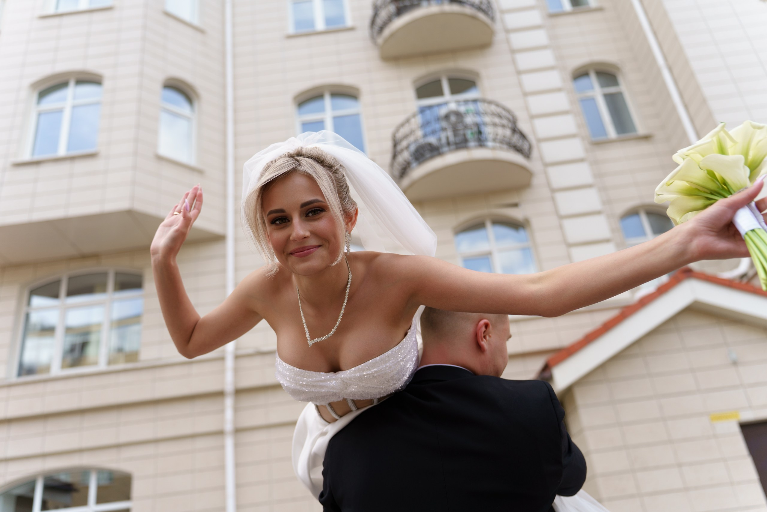 Жених и невеста на свадебной фотосессии: невеста в изысканном белом платье сидит на плечах жениха, машет рукой и держит букет светло-зелёных цветов. На фоне — здание с балконами | творческий свадебный фотограф Екатеринбург