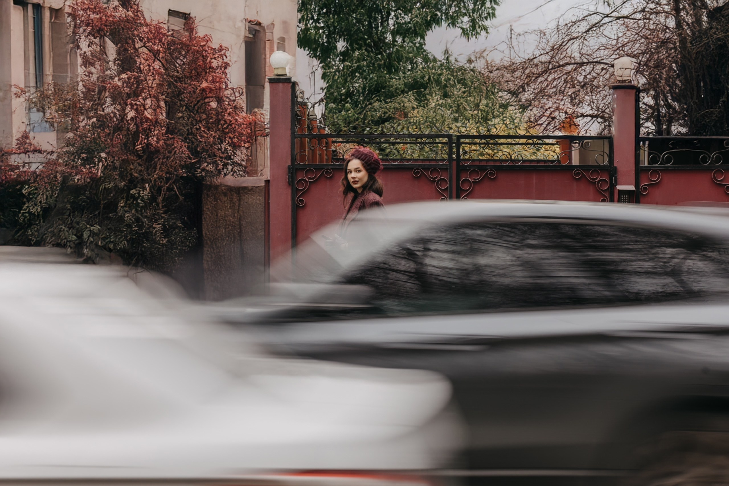 Девушка стоит у кованых ворот на фоне дома, мимо мчатся машины. Атмосфера немного пасмурная, городская суета.
