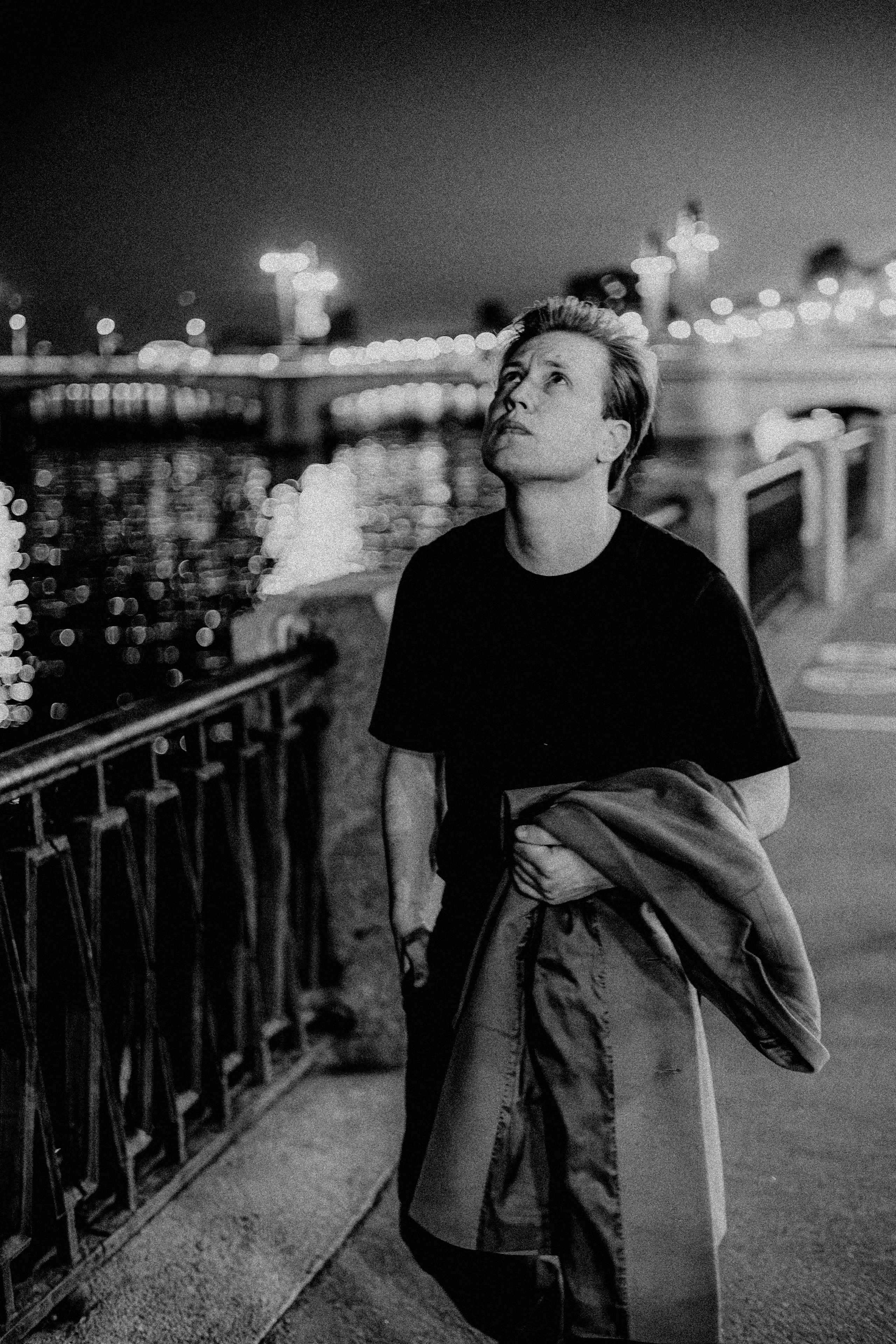 Молодой человек в черной футболке с пальто в руке идёт по набережной реки.