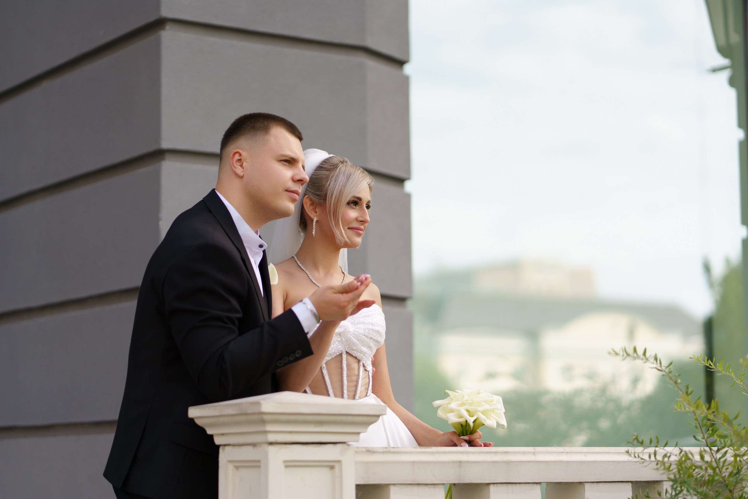 Невеста в белом платье с открытыми плечами и фатой и жених в чёрном костюме запечатлены в момент свадебной фотосессии. На заднем плане — серая архитектурная стена | свадебный альбом Екатеринбург