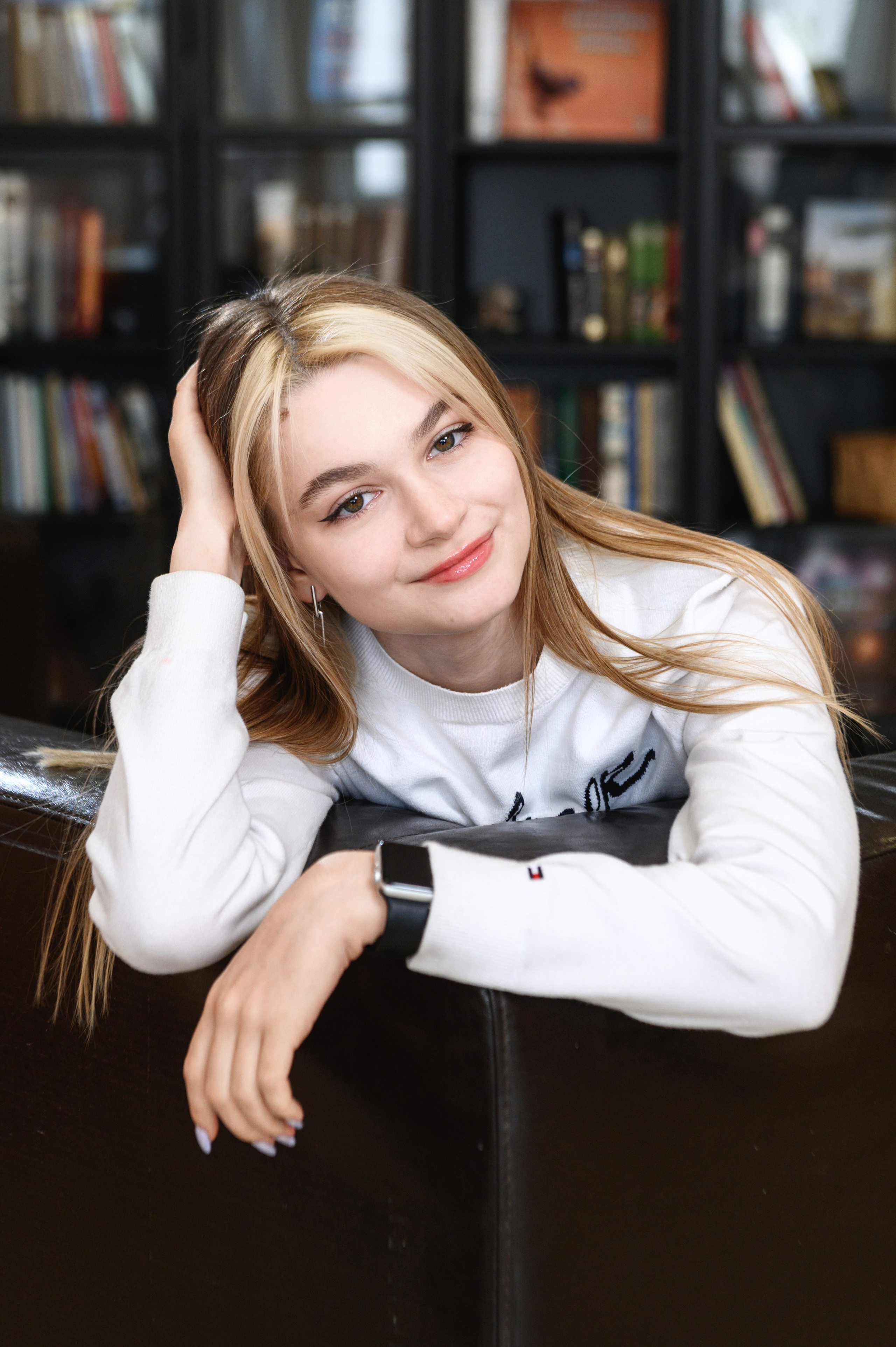Старшеклассница в светлом свитере позирует у стеллажей, опираясь рукой на подлокотник кресла.