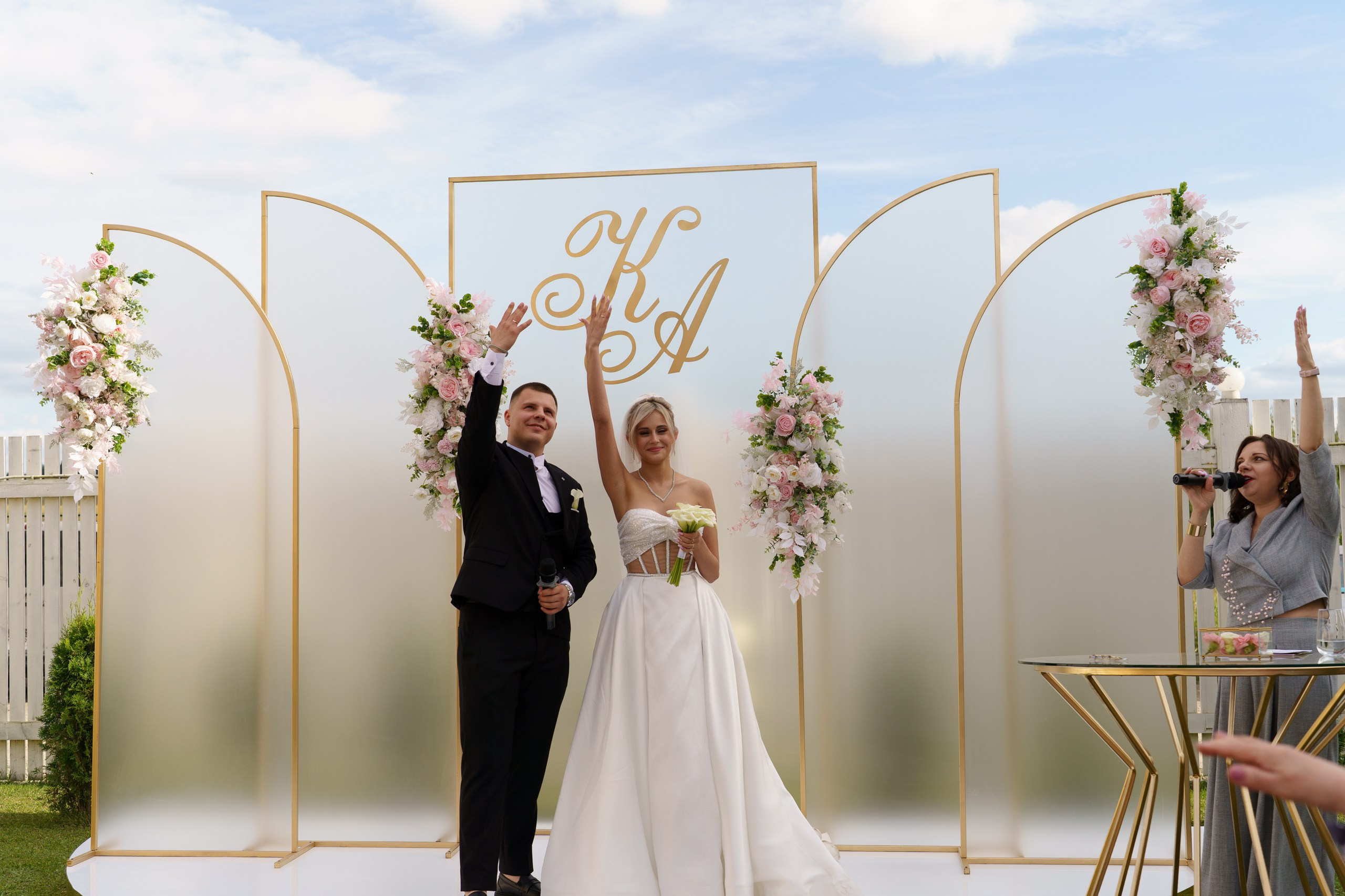 Молодожёны на открытой свадебной церемонии приветствуют гостей на элегантной бело‑золотой арке с цветочными композициями и инициалами, пока ведущая с микрофоном объявляет торжественный момент | фотограф для выездного банкета Екатеринбург