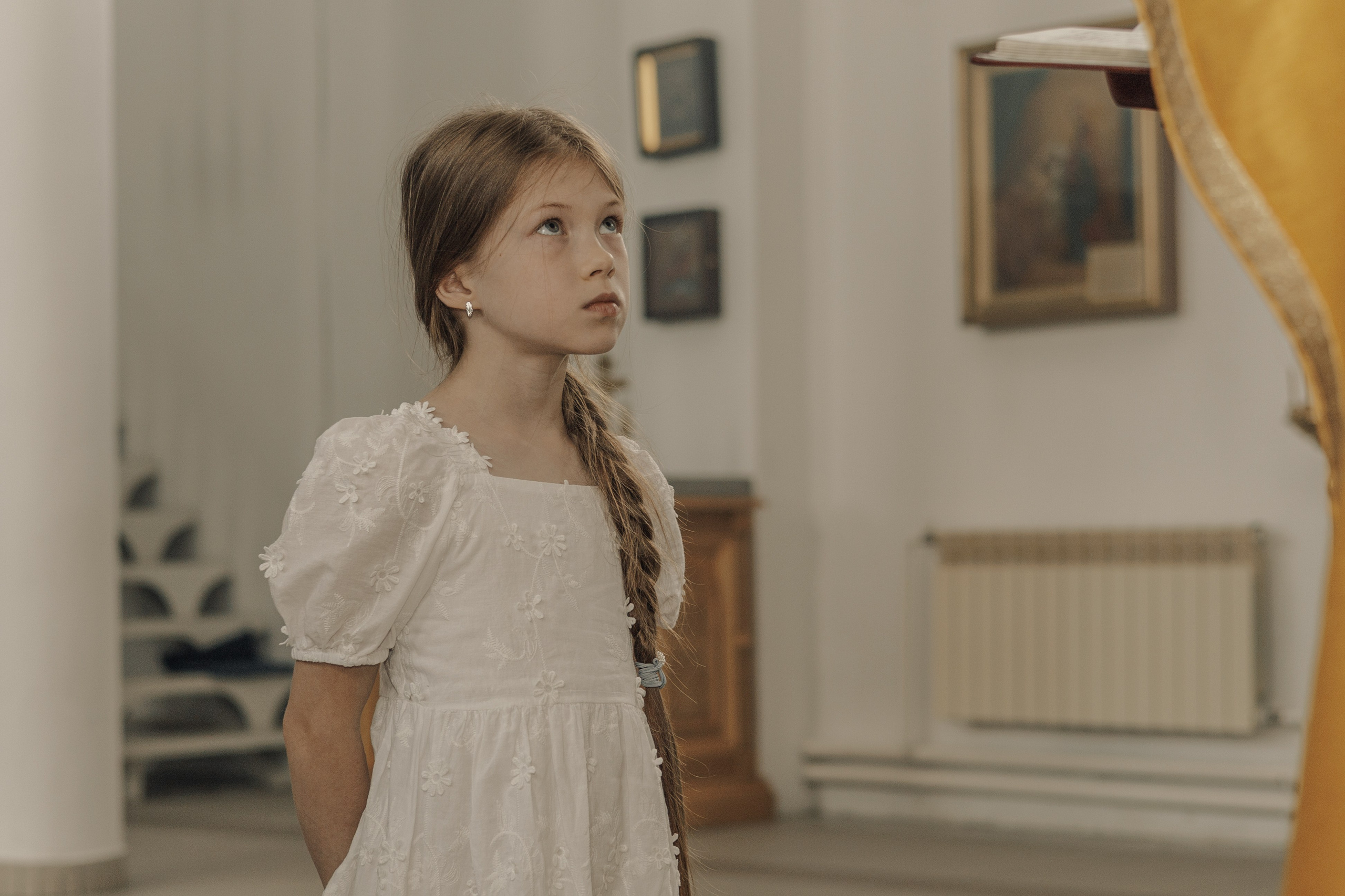 Девочка в белом платье с заплетенными в косу волосами смотрит вверх в мягко освещенном храме, украшенном иконами, передающими чувство благоговения и любопытства.