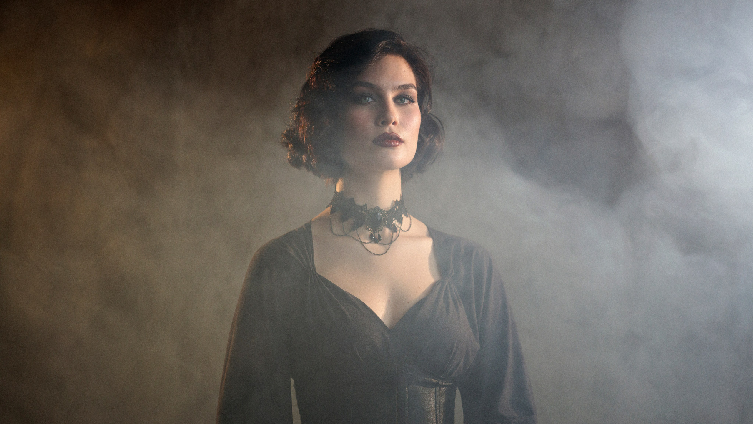 Портрет девушки в анфас, средний план, глаза ярко-зелёного цвета, тёмные волосы и губы накрашены ярко-терракотовой помадой, волосы до плеч, уложены в ретро укладку. Платье чёрного цвета, девушка стоит в дыму.