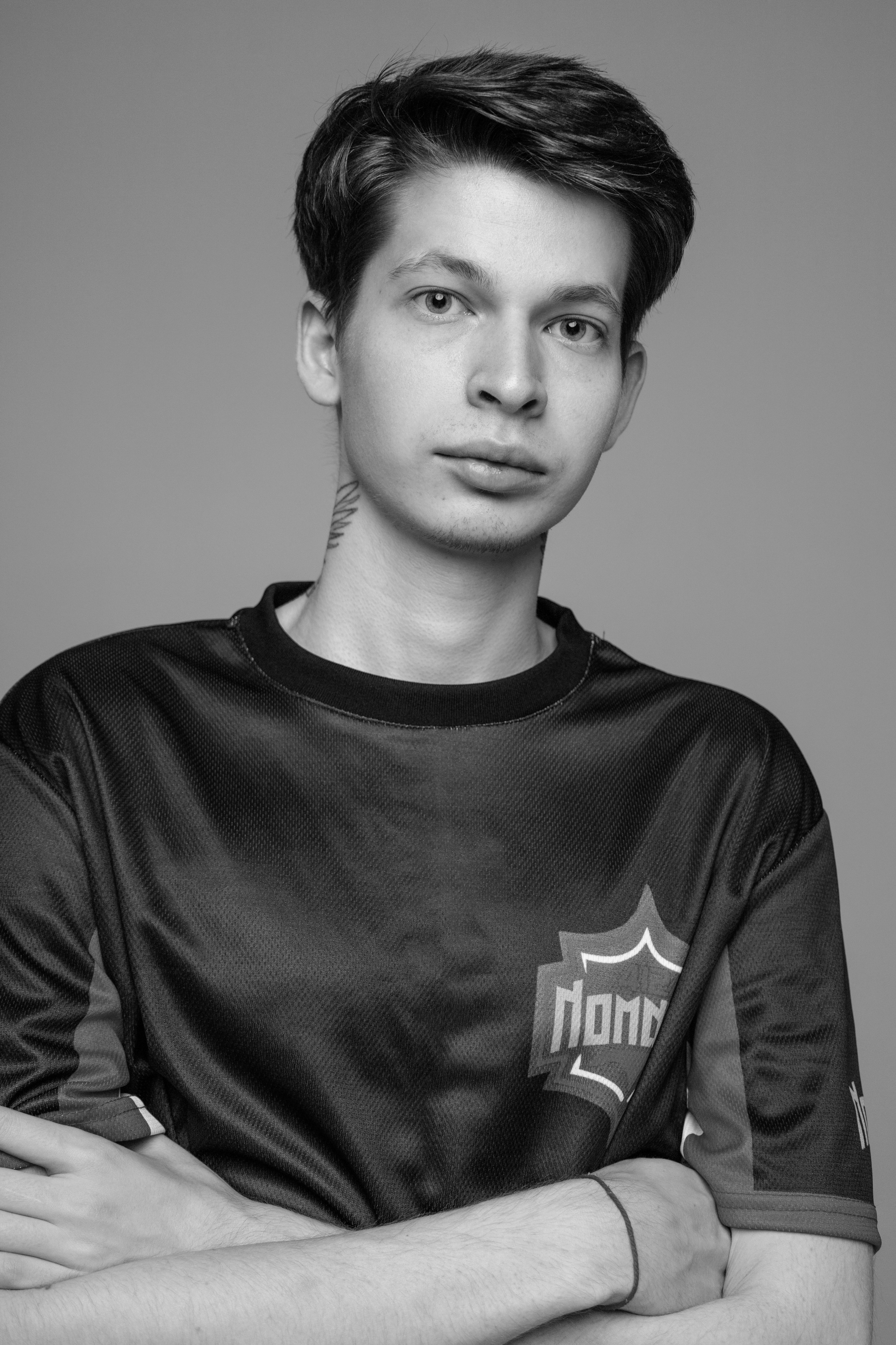 Молодой человек с короткой стрижкой смотрит в камеру, скрестив руки на груди, одетый в черную спортивную майку с узорчатым логотипом. Атмосфера нейтральная и спокойная.