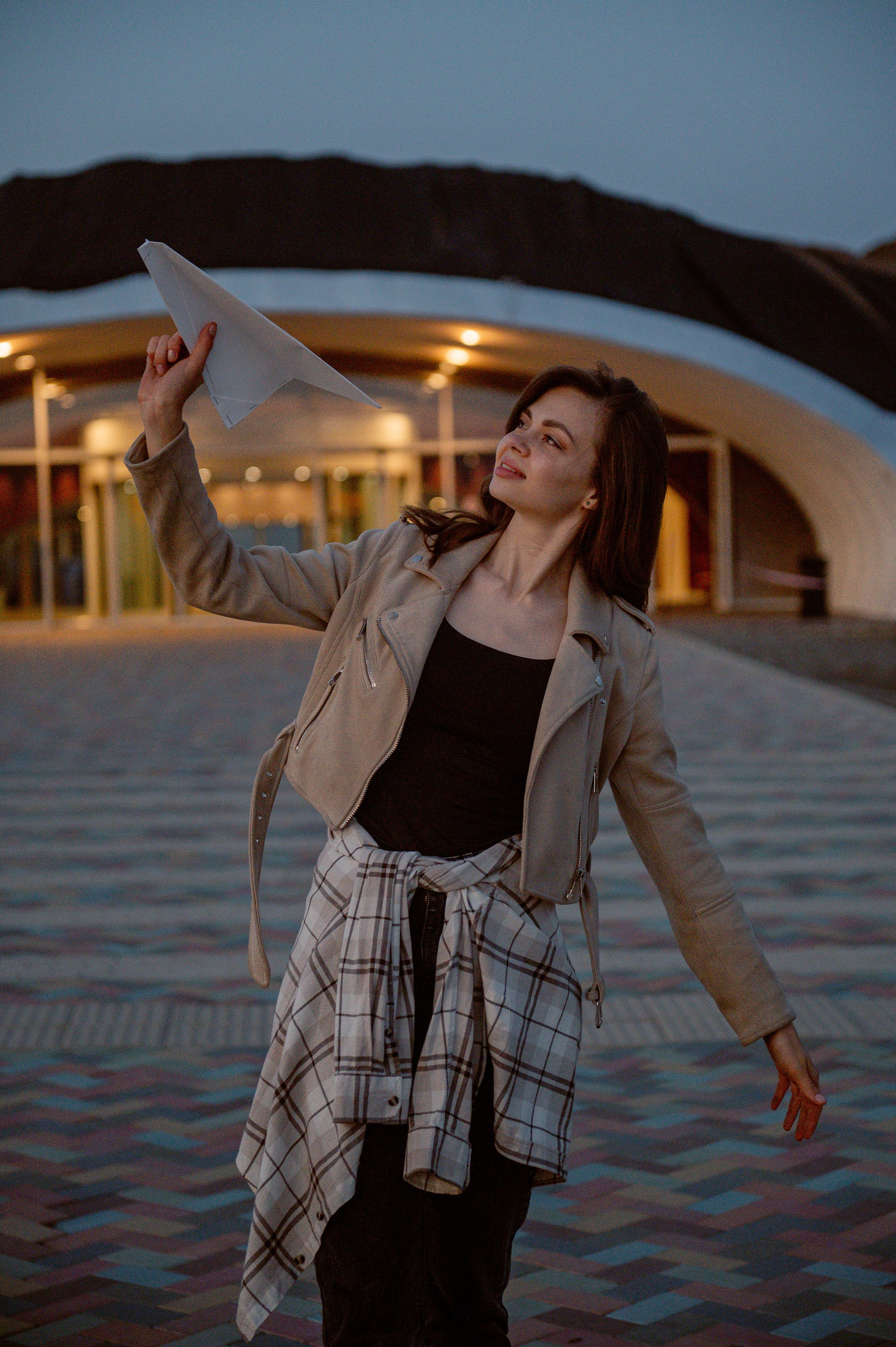 Девушка в светлой куртке и клетчатой рубашке держит бумажный самолётик. На фоне — современное здание, сумерки, атмосфера лёгкая и игривая.