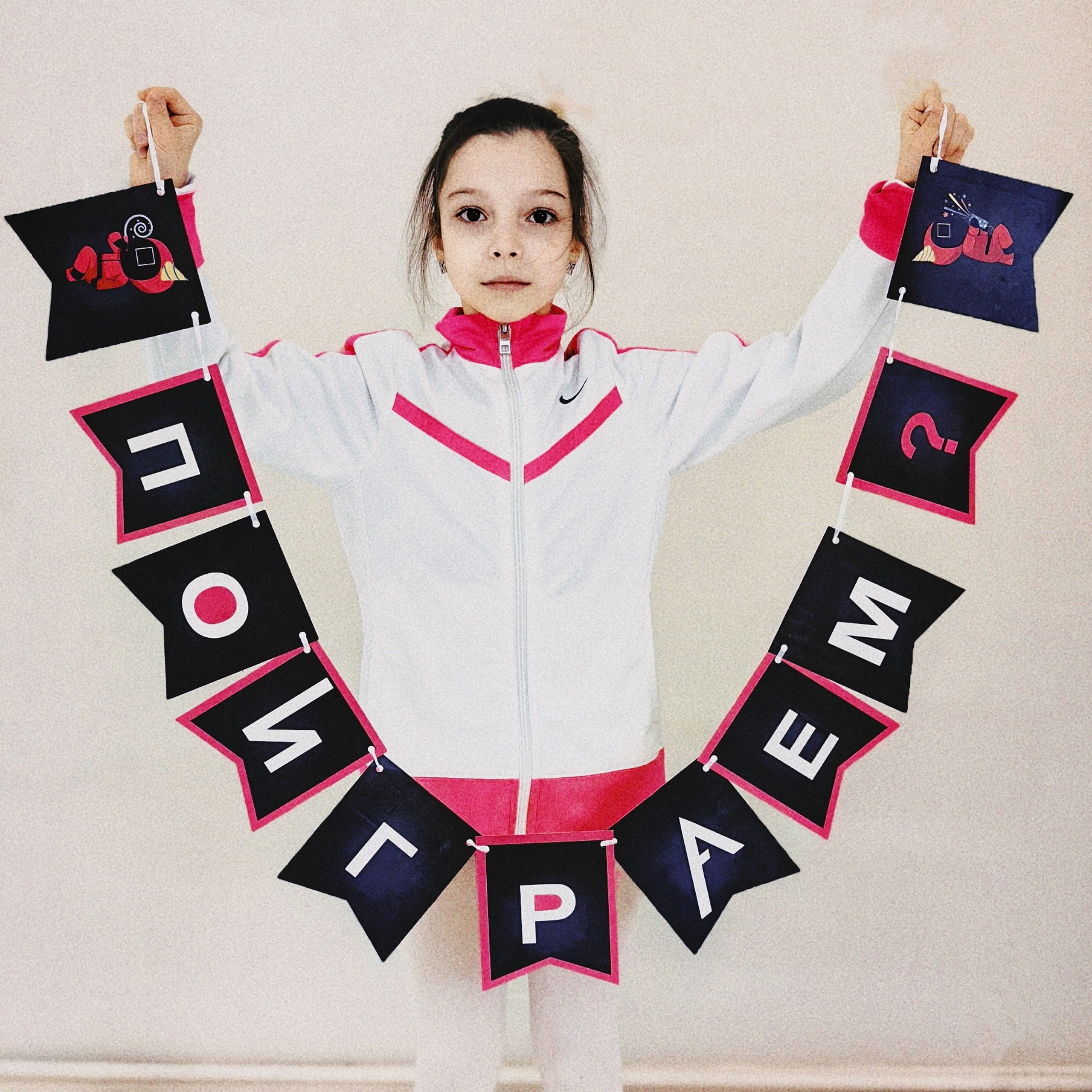 Девочка демонстрирует вывеску "Поиграем" как приглашение принять участие в игре в стиле "Игры в кальмара".