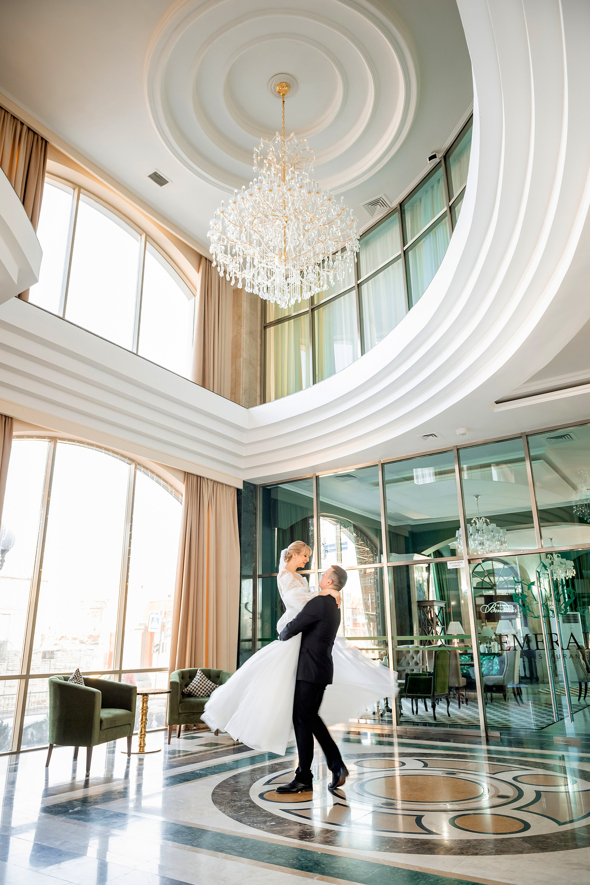Свадебная пара в элегантном интерьере: жених поднимает невесту на руках в просторном зале с высокими окнами, мраморным полом и большим хрустальным люстрой.