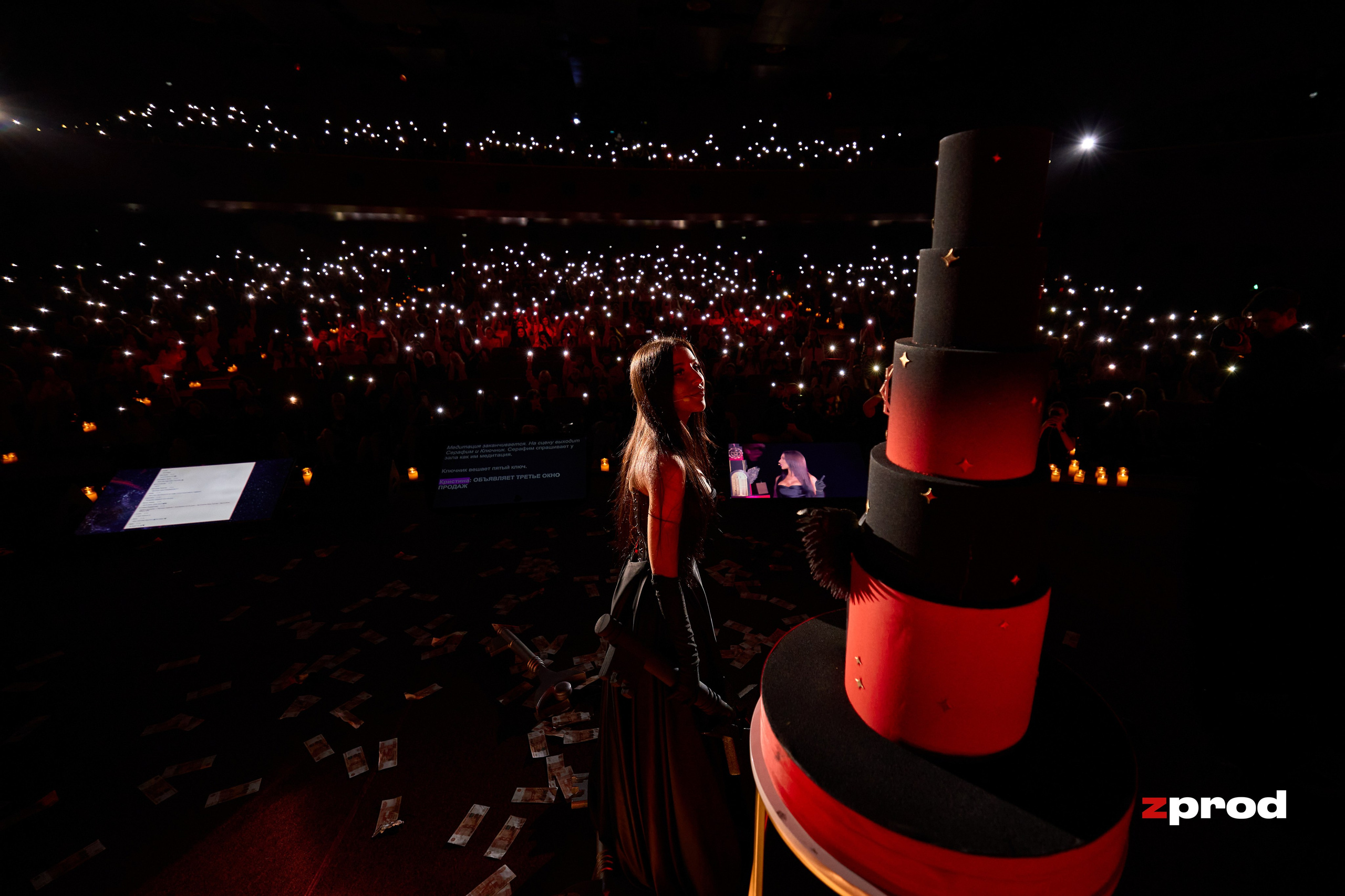 Кадр с крупного мероприятия в кинотеатре 'ОКТЯБРЬ': на сцене стоит Кристина Егиазарова, ведущая презентацию в области инфобизнеса и цифровой психологии. В фокусе — многослойный торт, окружённый огоньками мобильных телефонов в руках зрителей. Атмосфера торжественности и вдохновения на фоне яркого света и эффектного оформления