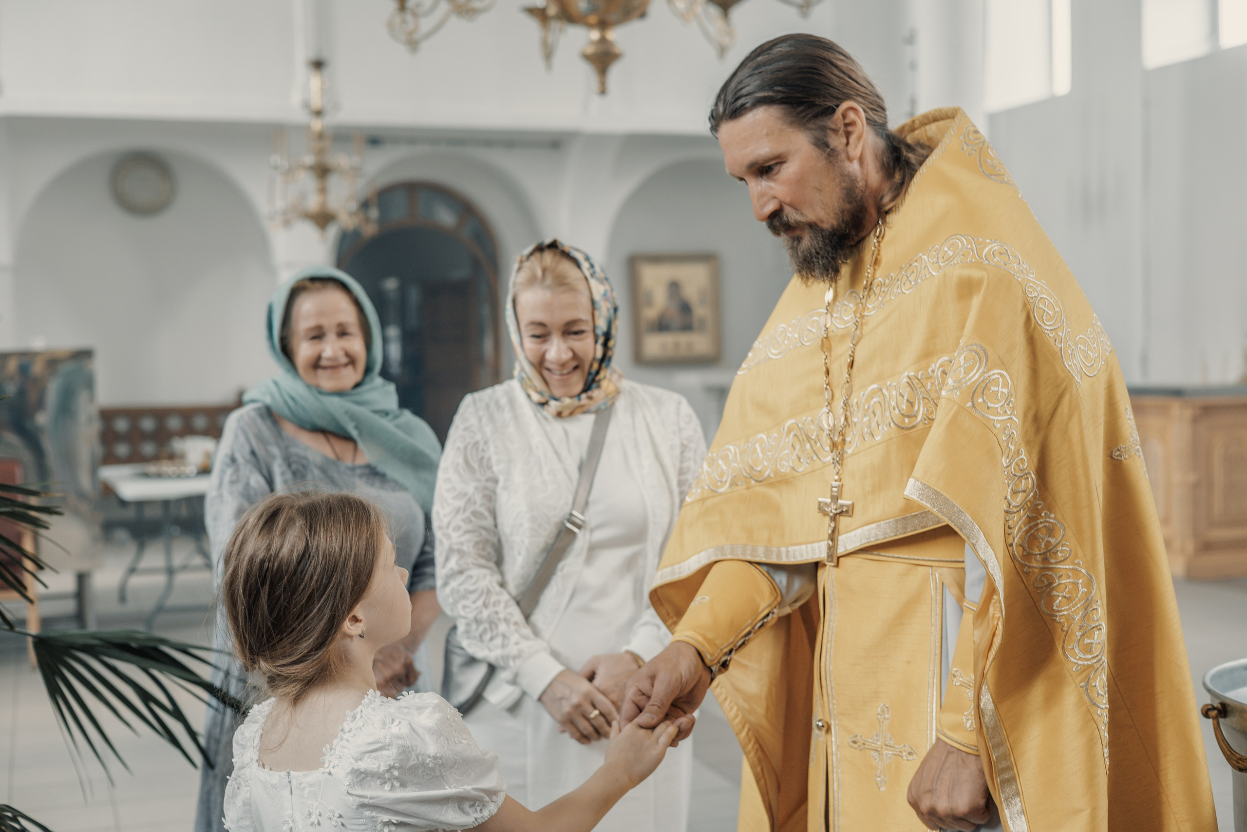 Священник в золотистой рясе нежно держит за руку девочку. Две женщины в платках стоят неподалеку, тепло улыбаясь в церковной обстановке.