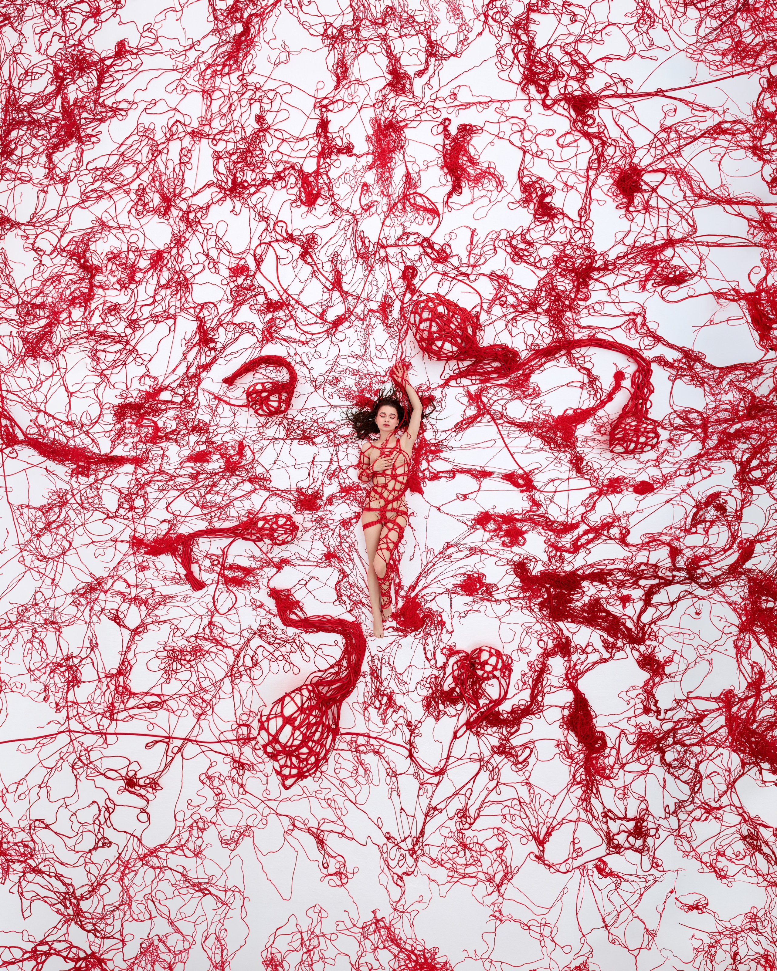 Творческий проект: на фотографии изображена женщина, лежащая на белом фоне, обвязанная красной нитью, которая расползается по всей площади снимка в виде сложного узора. Нити образуют абстрактные формы, создавая динамичную и визуально насыщенную композицию, подчеркивающую контраст между чёткостью фигуры и хаосом красных линий
