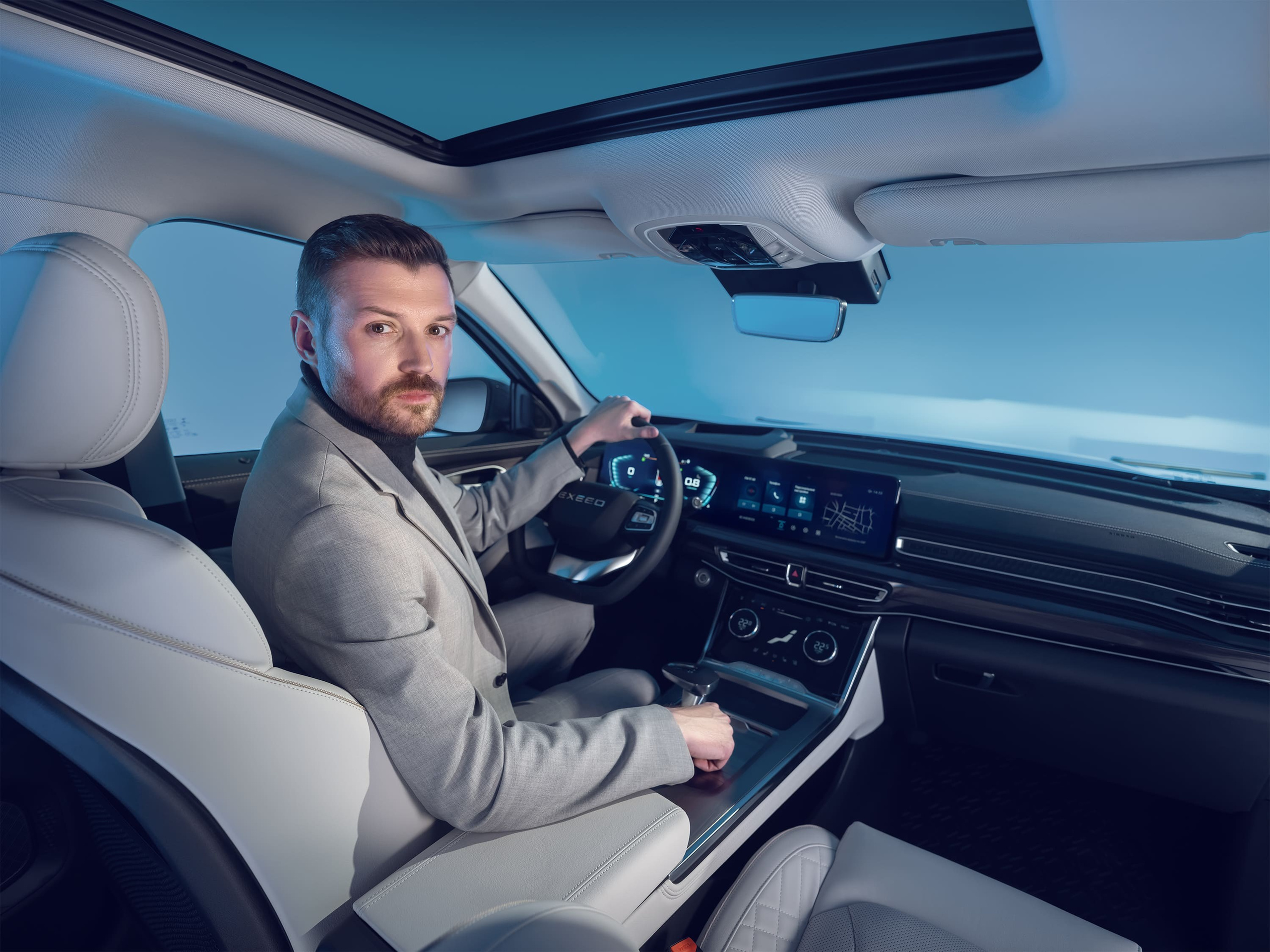 Мужчина с короткой щетиной в сером пиджаке сидит за рулем автомобиля Exeed