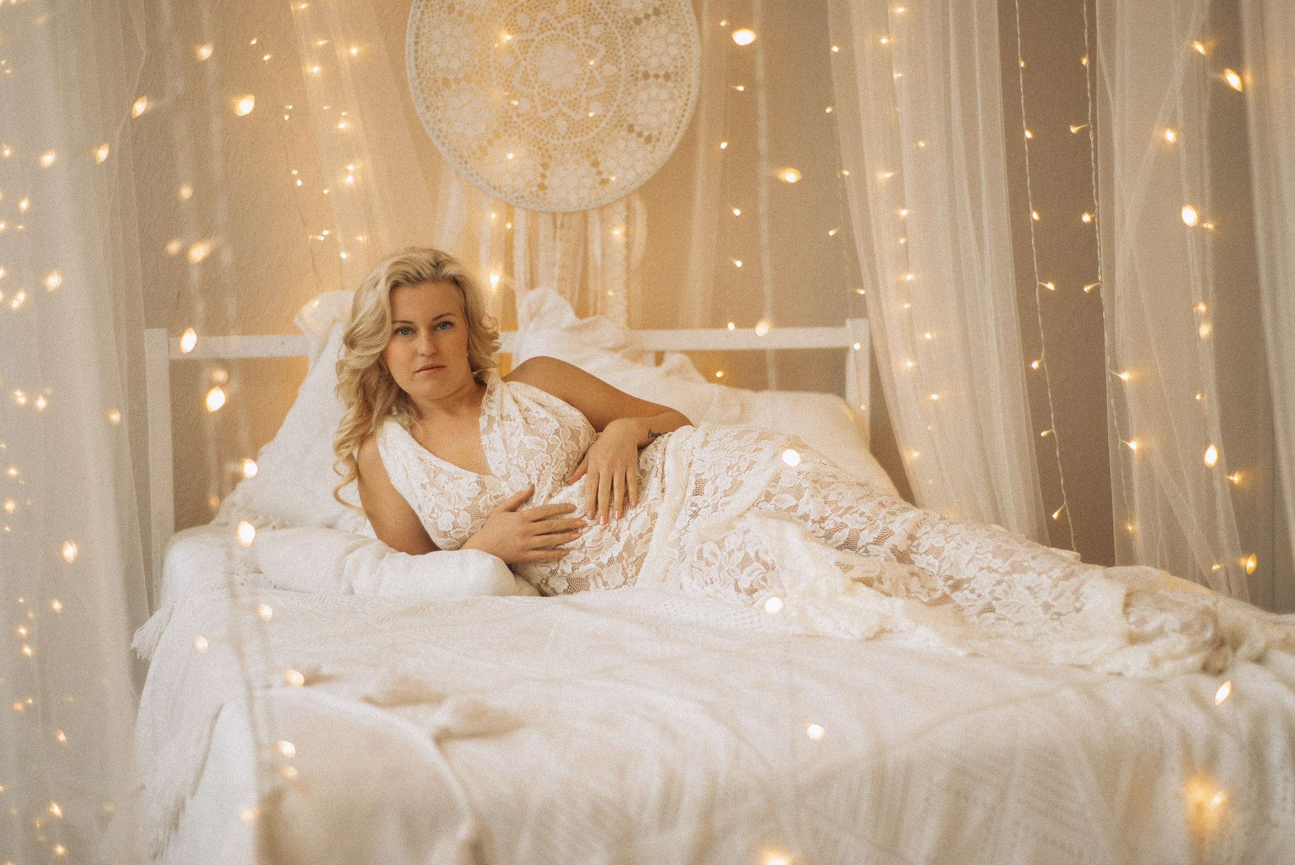 Натали Каролин лежит на белой кровати в кружевном платье. Атмосфера бохо-студии с ловцом снов и теплым светом гирлянд. Профессиональная женская фотосессия в Новосибирске