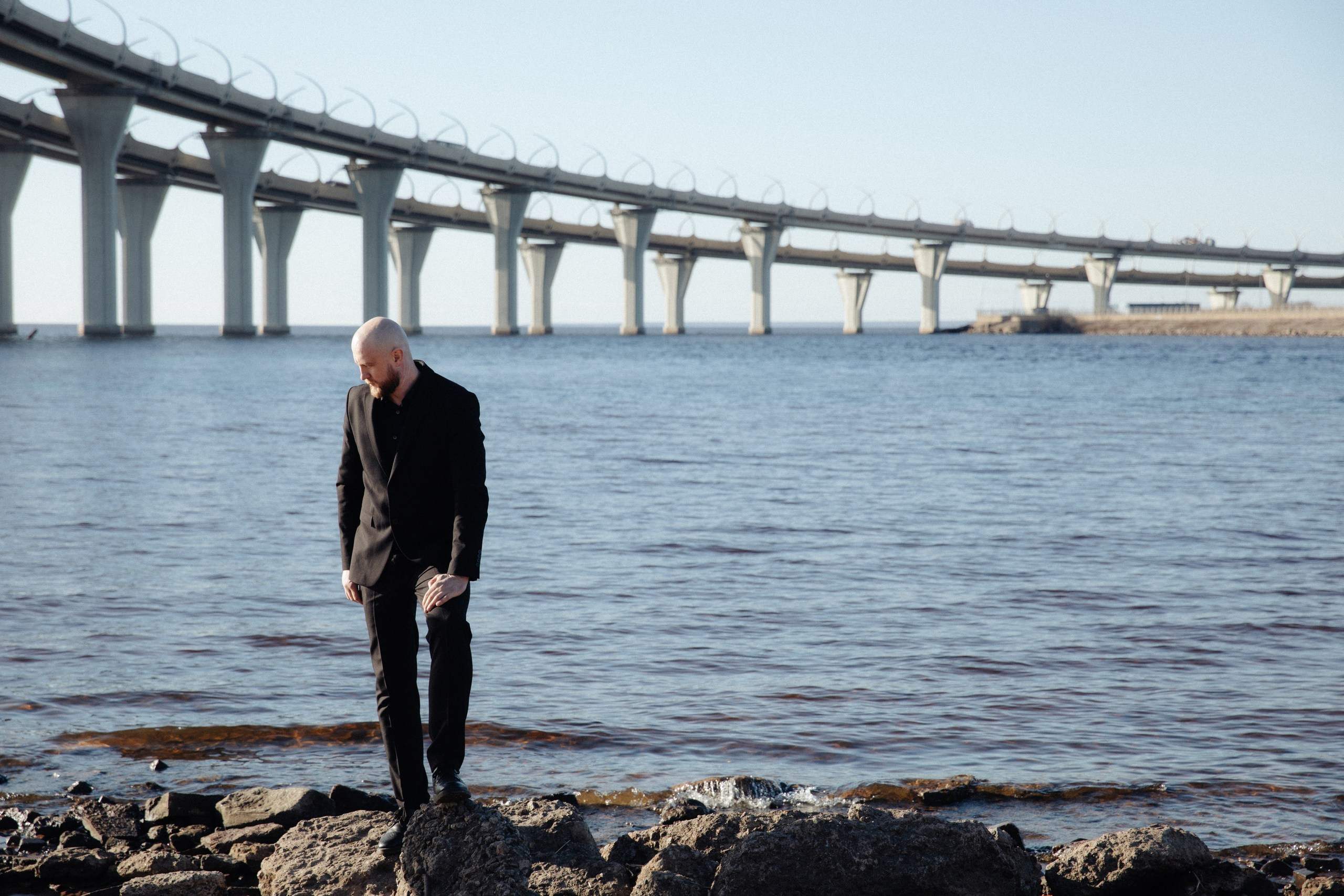 мужчина в черном костюме стоит на мостовой у воды, современный портрет в Санкт-Петербурге.