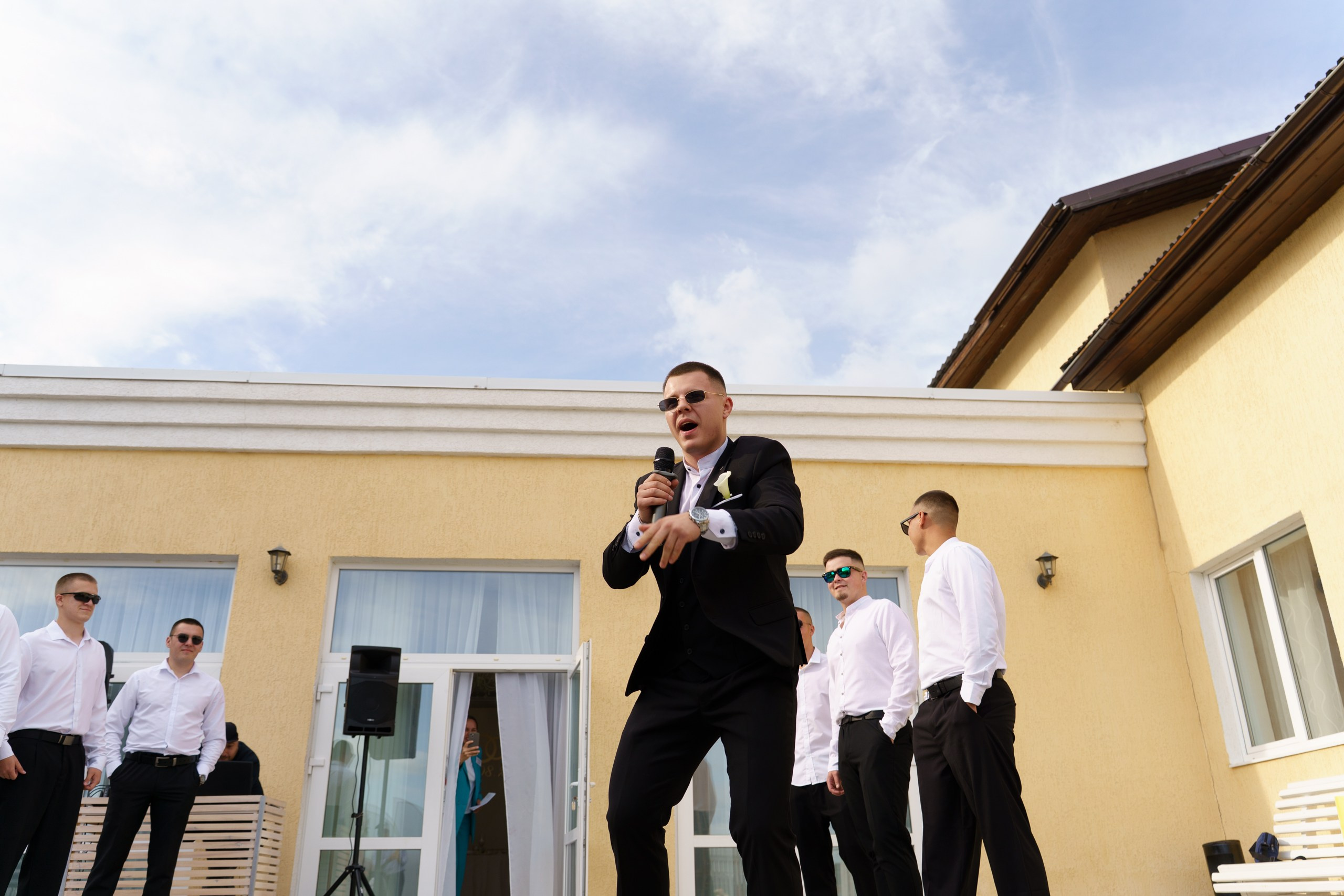 Праздник в разгаре: стильный жених в солнцезащитных очках и смокинге ведёт мероприятие на открытом воздухе, а его команда в элегантных нарядах создаёт ощущение гармонии и торжества | свадебный репортаж Екатеринбург