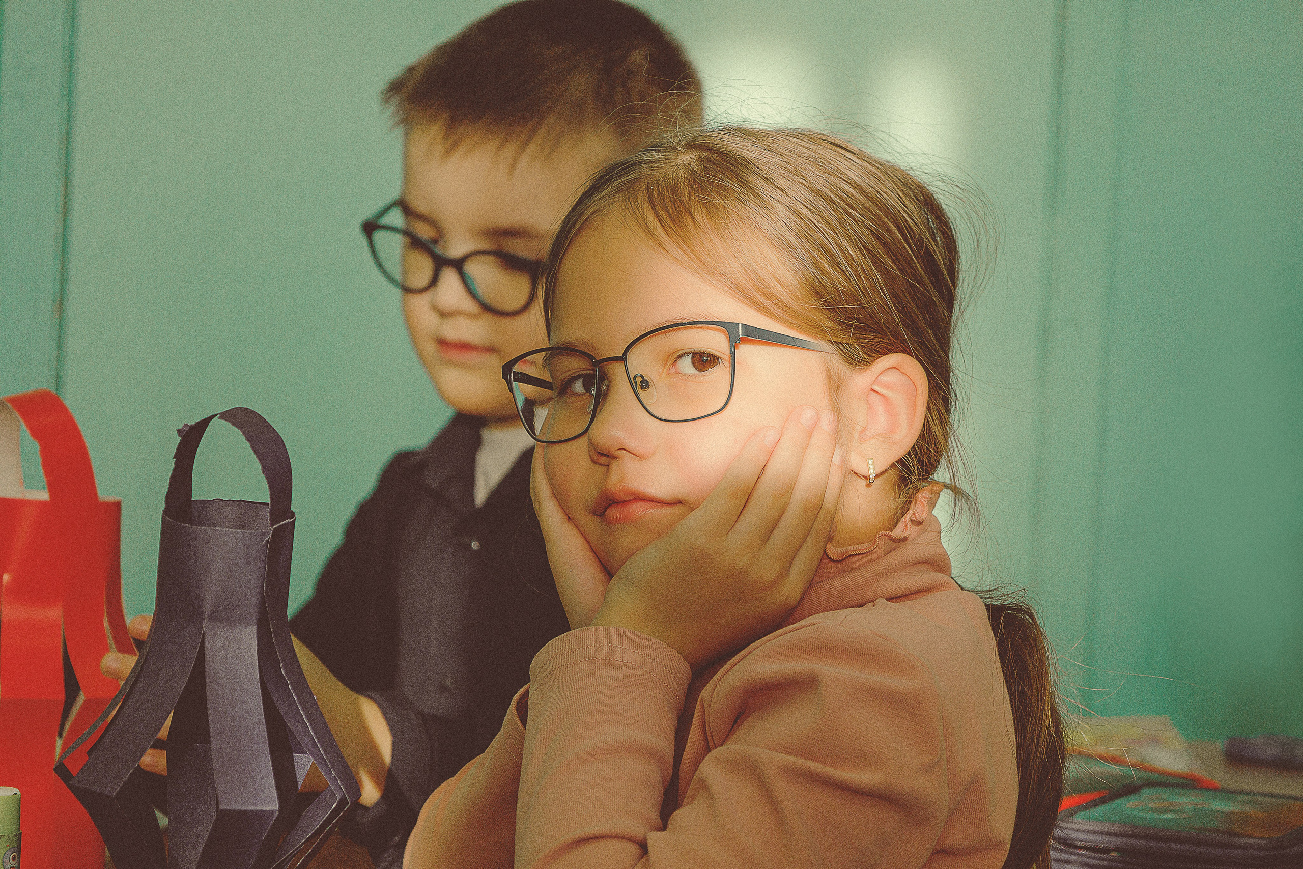 Двое детей в очках сидят за партой в школе и занимаются рукоделием. Девочка с задумчивым видом подпирает подбородок руками, а мальчик работает рядом с ней. Видны разноцветные поделки из бумаги.