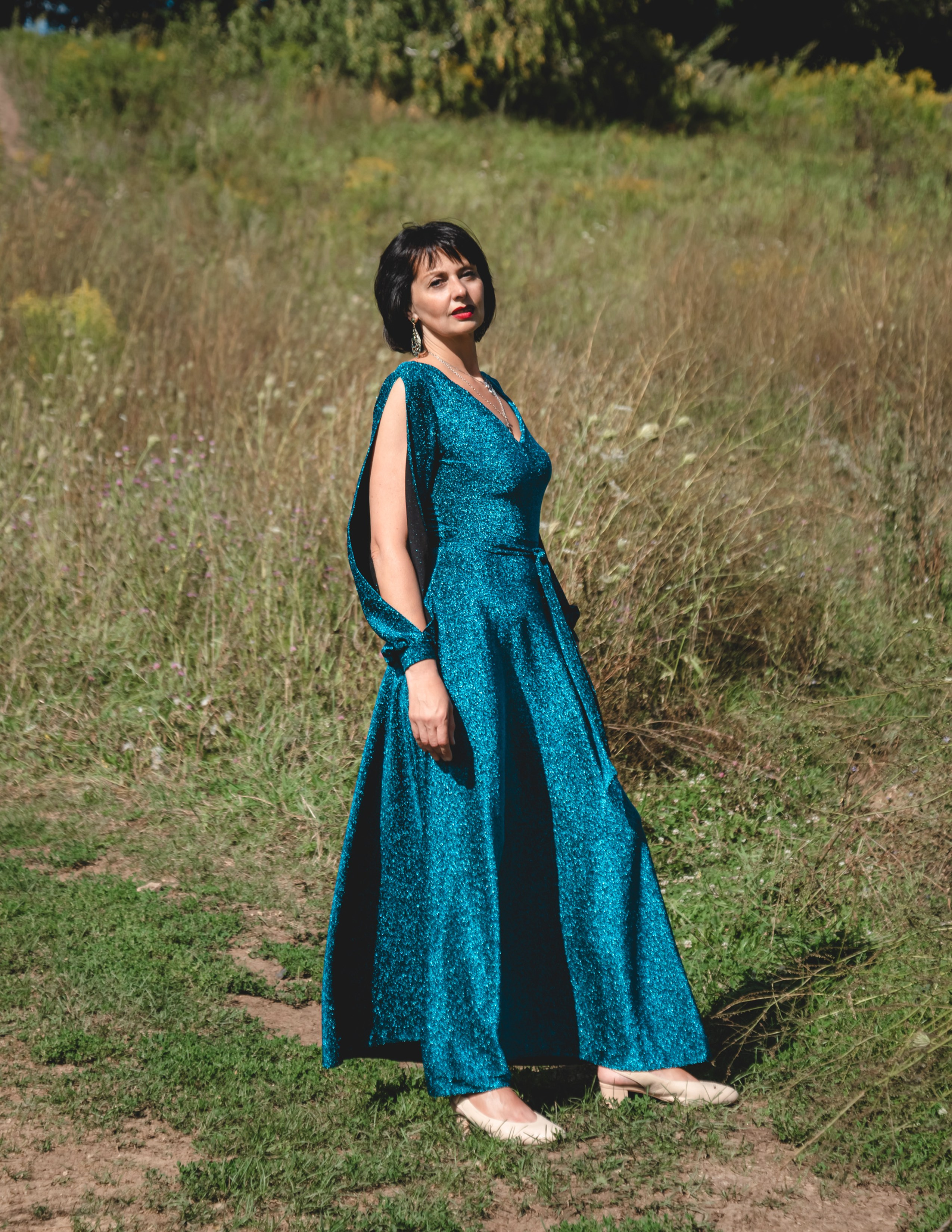 На фото женщина в изумрудном платье стоит в поле.