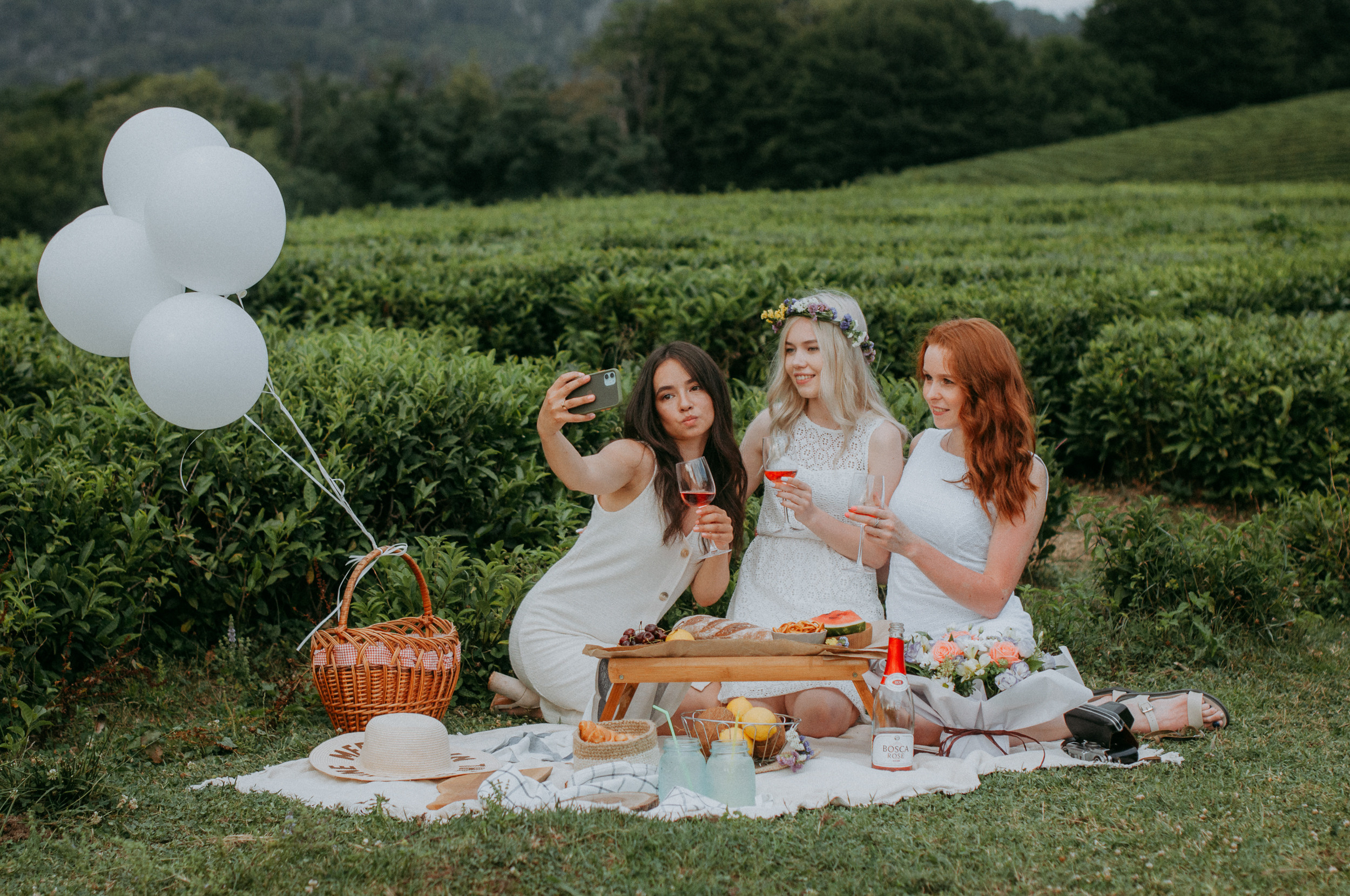 Три девушки фотографируются на телефон, делают селфи с бокалами на пикнике - девичнике на чайной плантации в Сочи