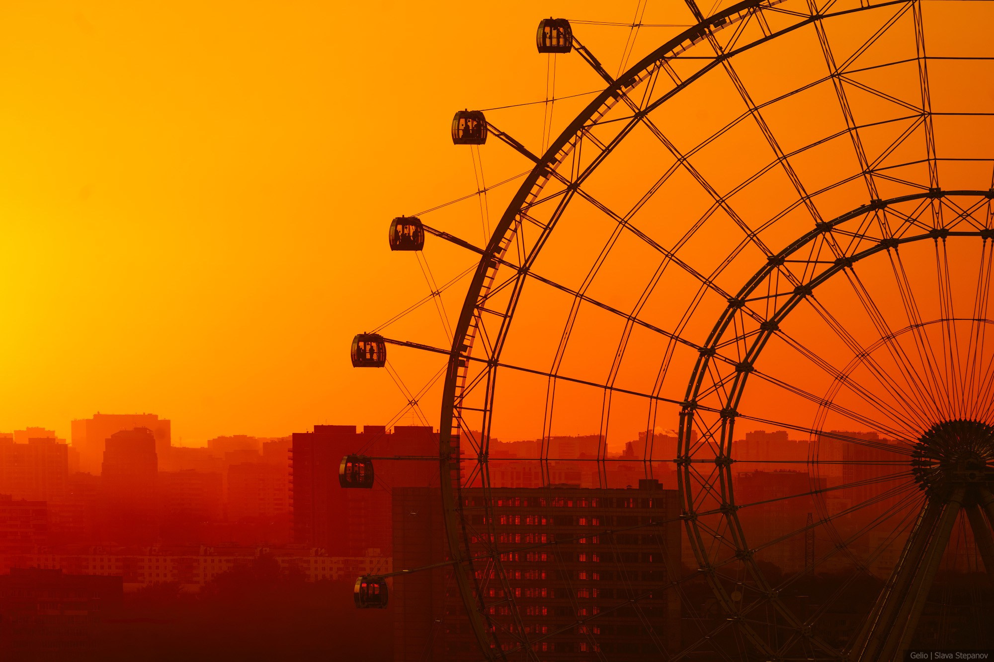 Close-up of the metal structures of the "Sun of Moscow" Ferris wheel against a bright yellow-orange sunset. Silhouettes of passenger cabins over the city. Photographer Slava Stepanov.