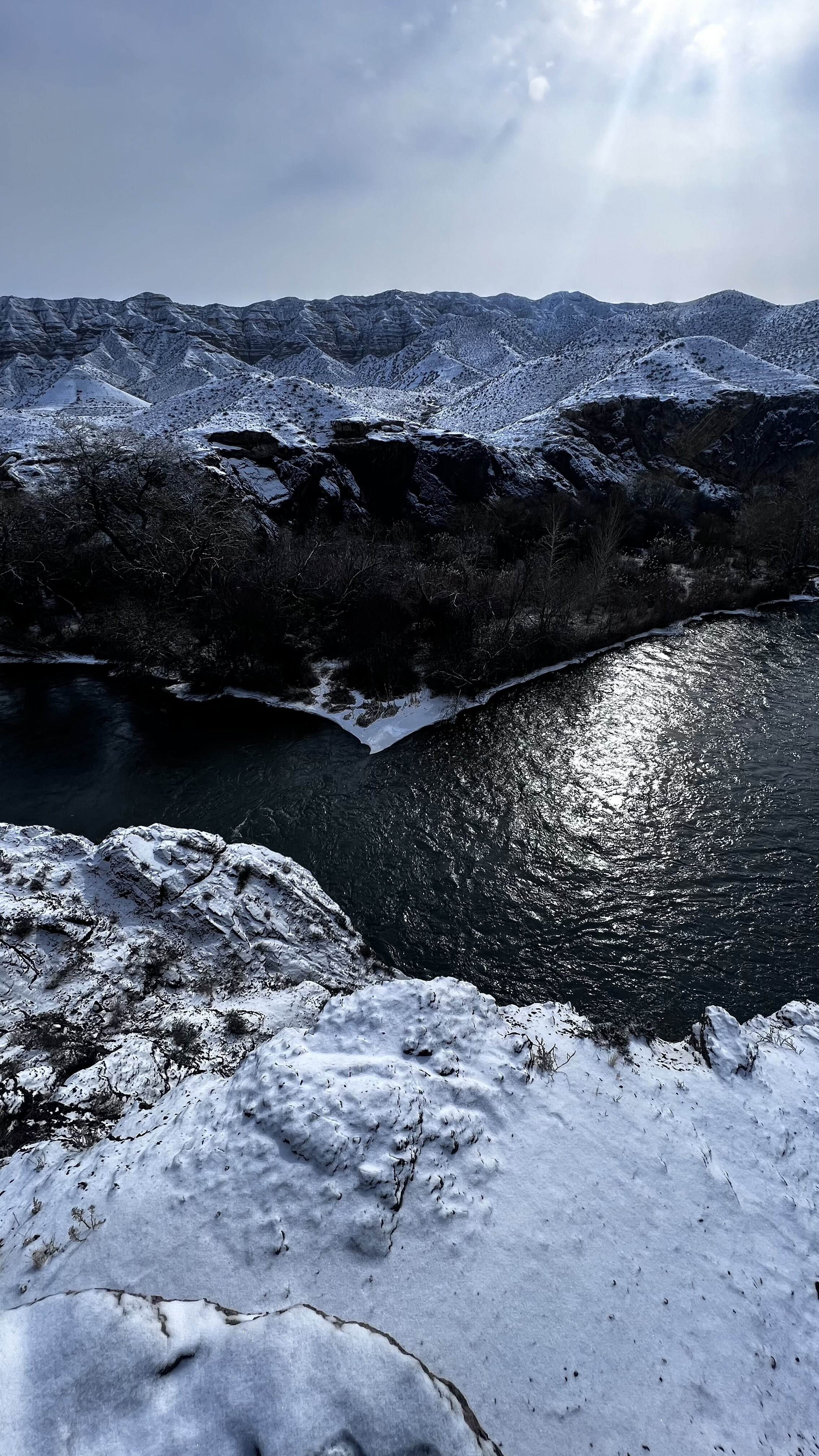 Зимний пейзаж Лунного каньона с видом на извилистую реку Чарын, окружённую заснеженными берегами. На переднем плане скалистая поверхность, покрытая снегом. Вода отражает приглушённый свет пасмурного неба. Вдали видны холмы, покрытые тонким слоем снега, придающим им мягкость и контраст с тёмными скалами.