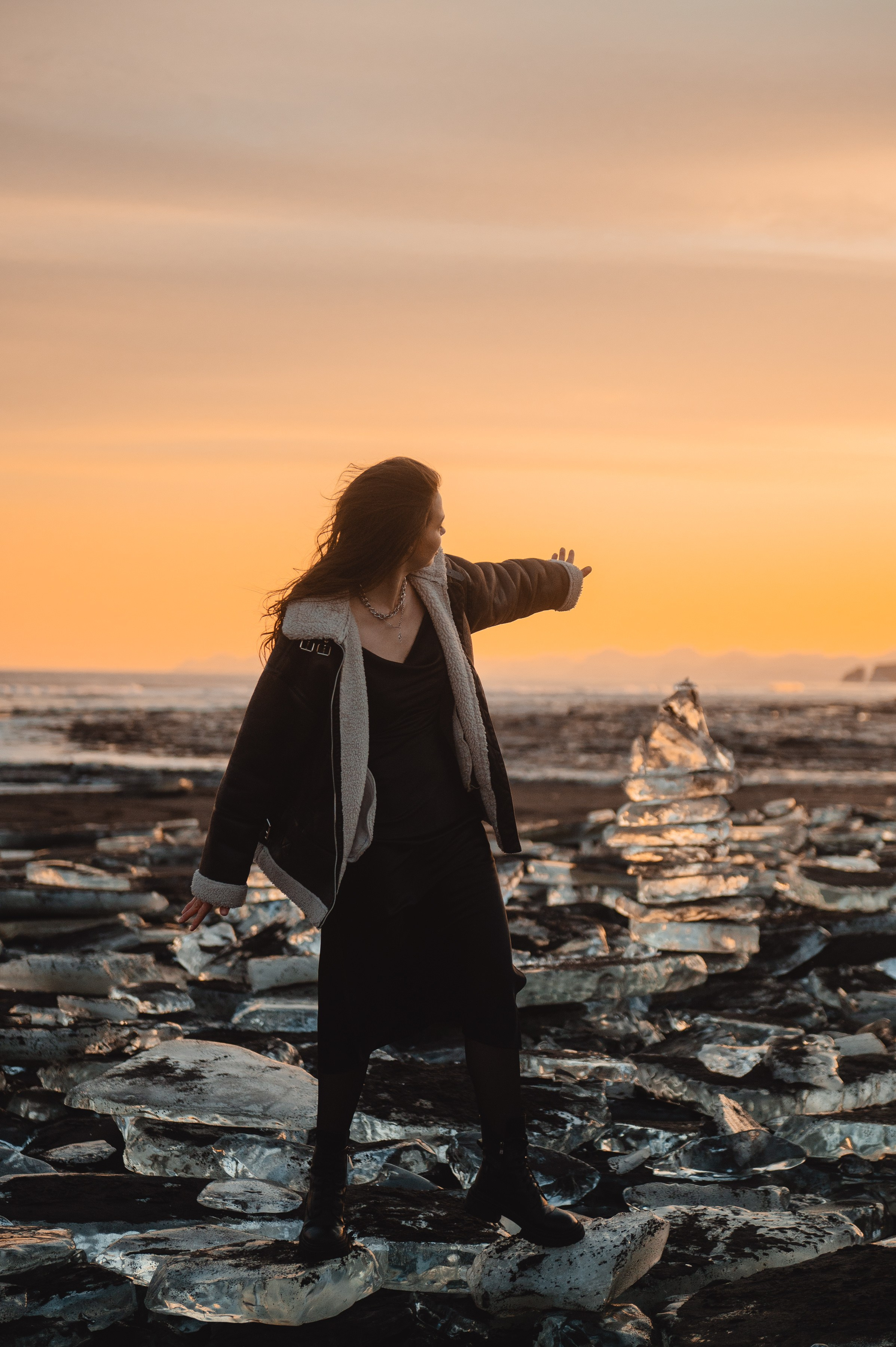 Девушка в чёрном платье и куртке стоит на ледяных глыбах у моря на закате. Атмосфера волшебная, немного холодная.