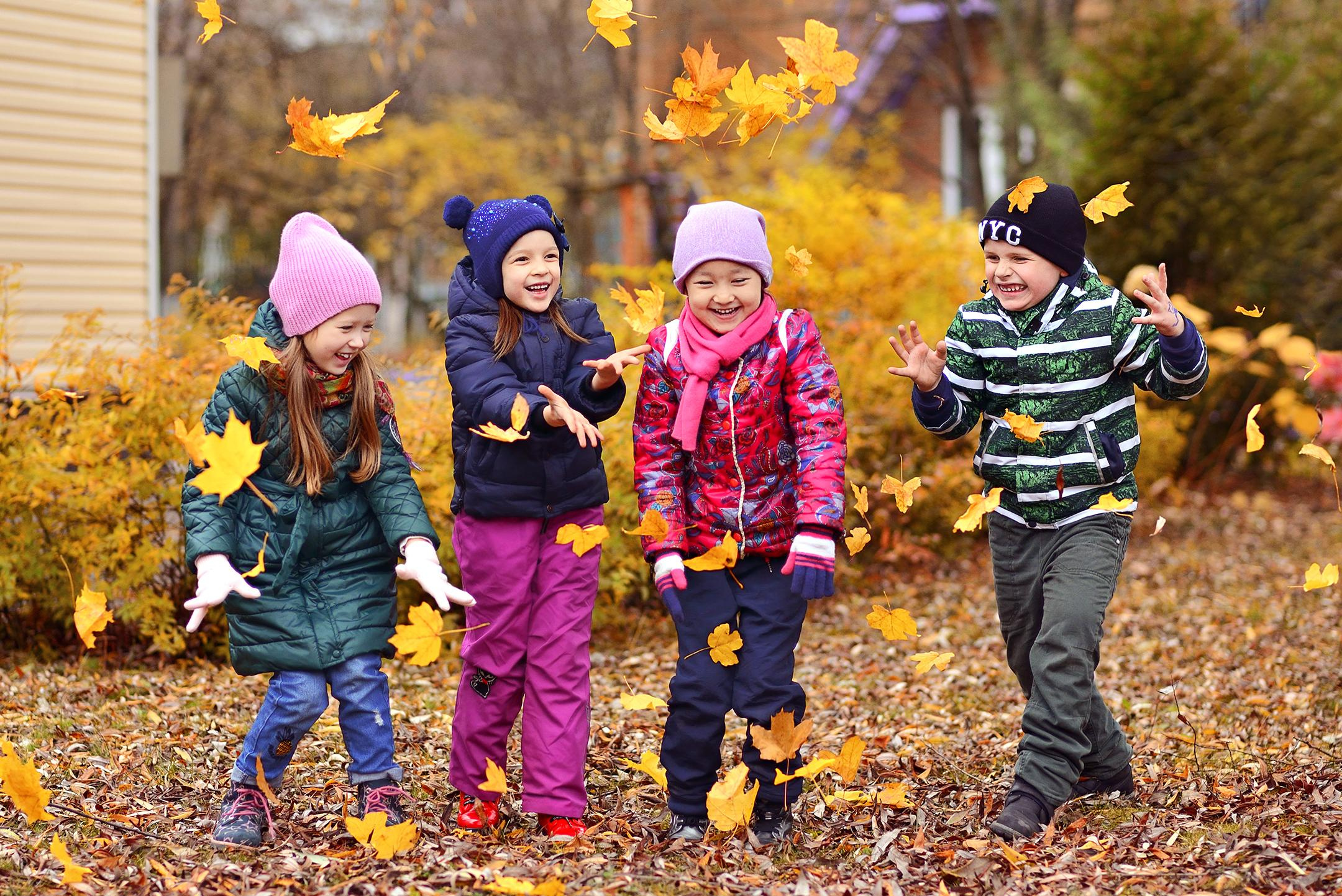 Четверо детей на прогулке в детском саду подкидывают листья вверх и смеются.