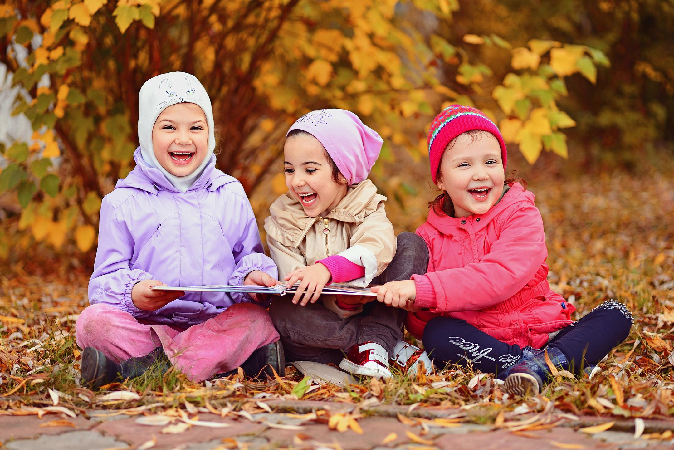 Трое детей читают книгу и смеются, сидя на опавшей листве во время прогулки в детском саду.
