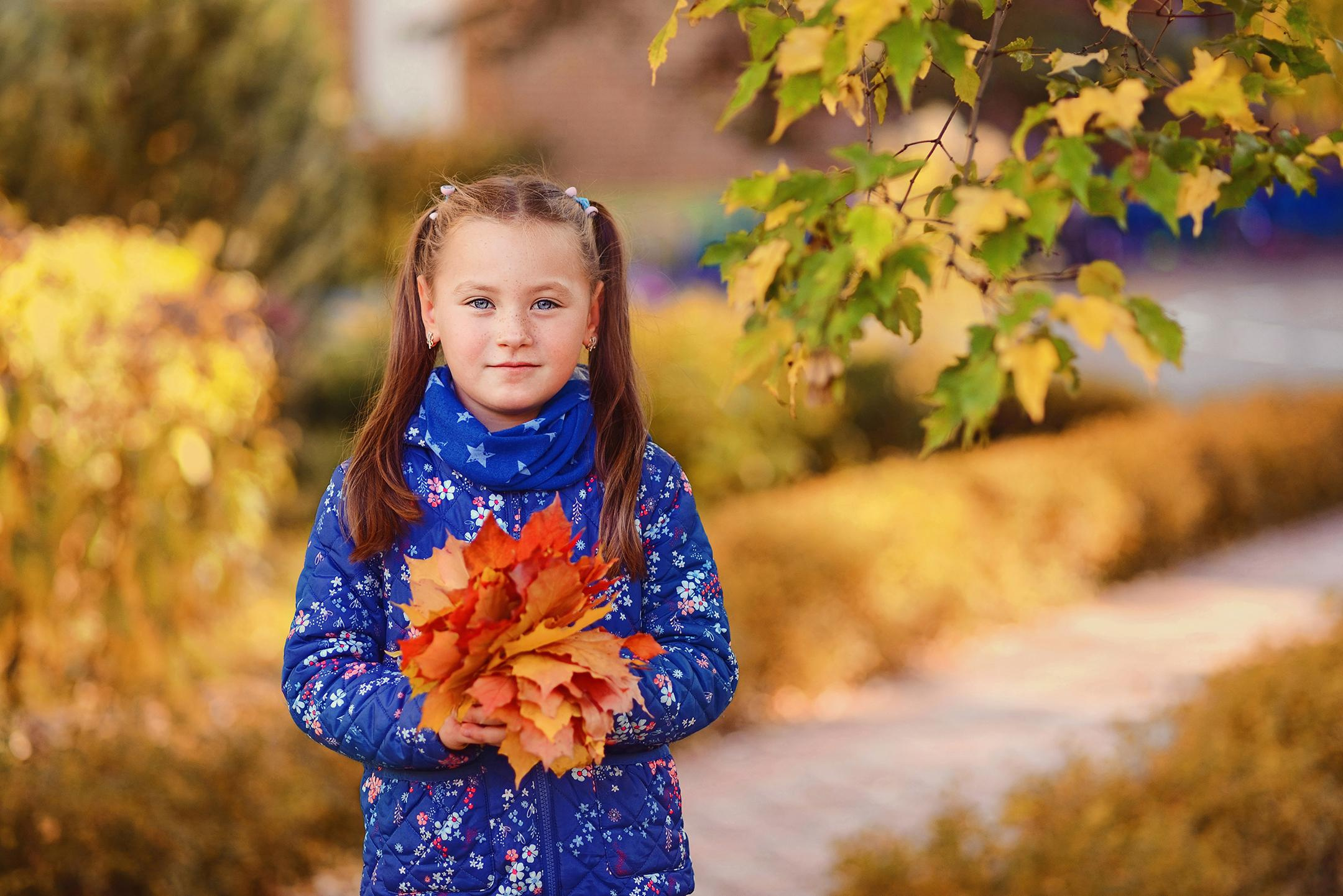 Девочка на фоне осеннего пейзажа держит букет из листьев.