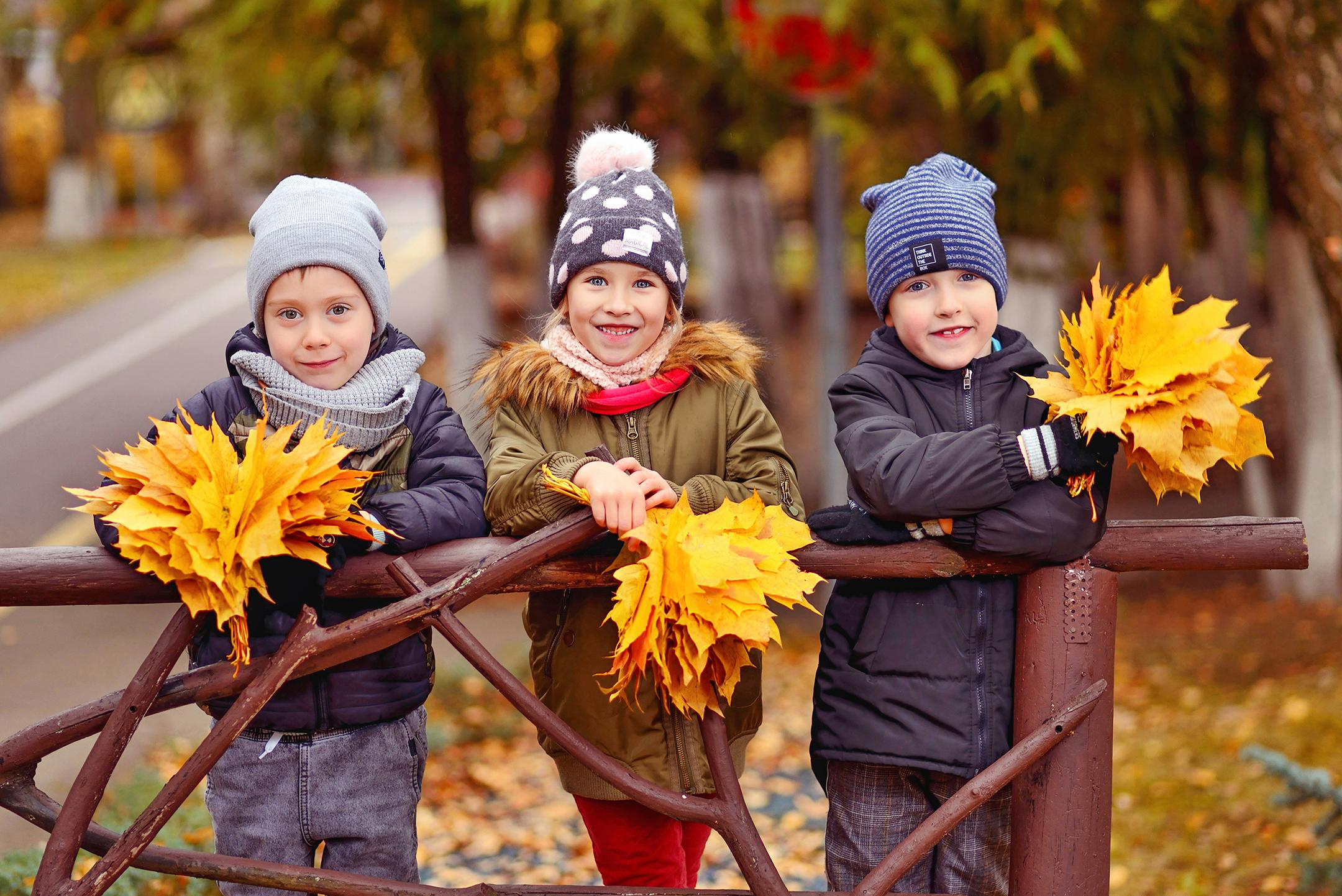 Трое детей на осенней прогулке в детском саду стоят на мостике с букетами из листьев.
