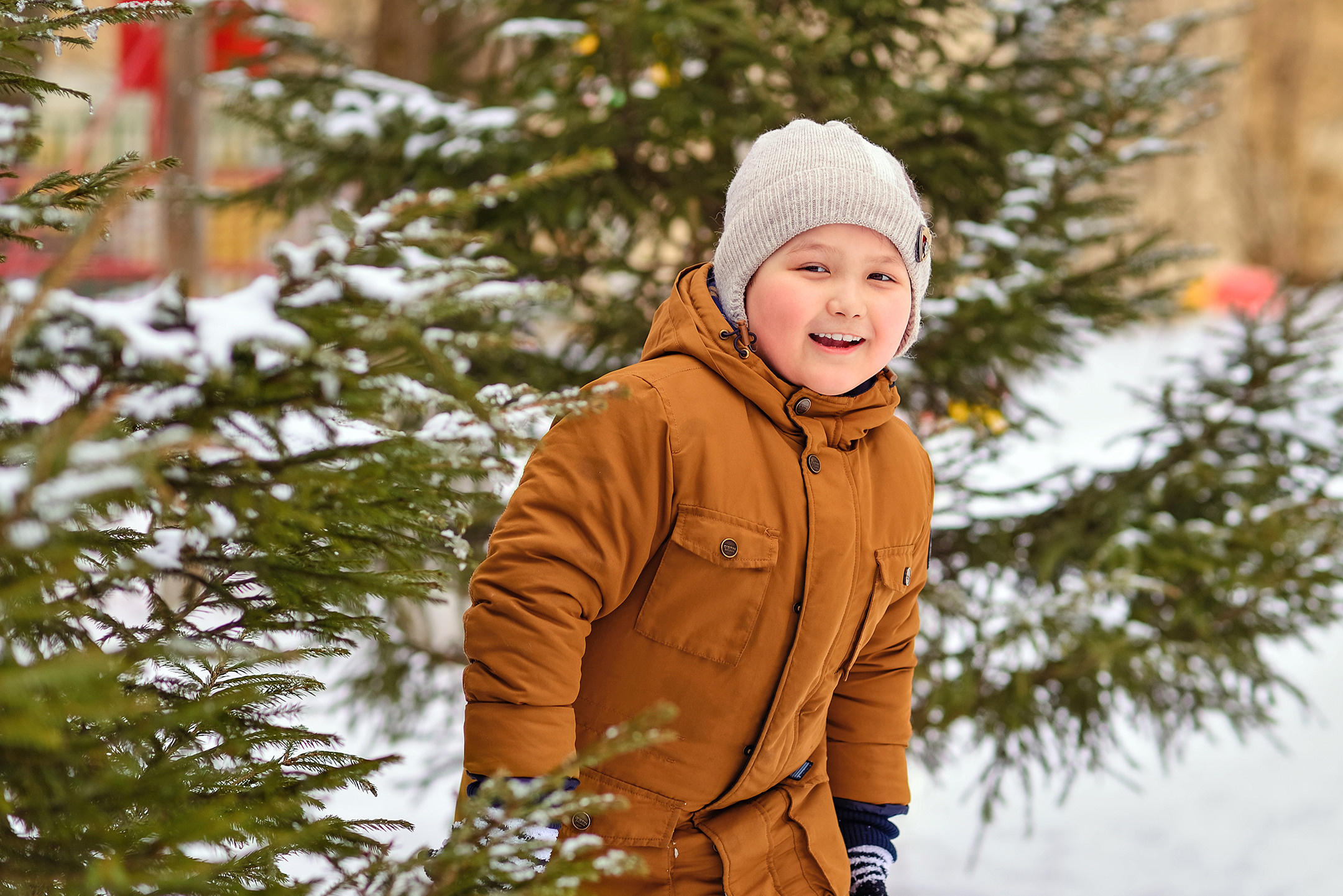 Мальчик на зимней прогулке в детском саду выглядывает из-за еловых веток.