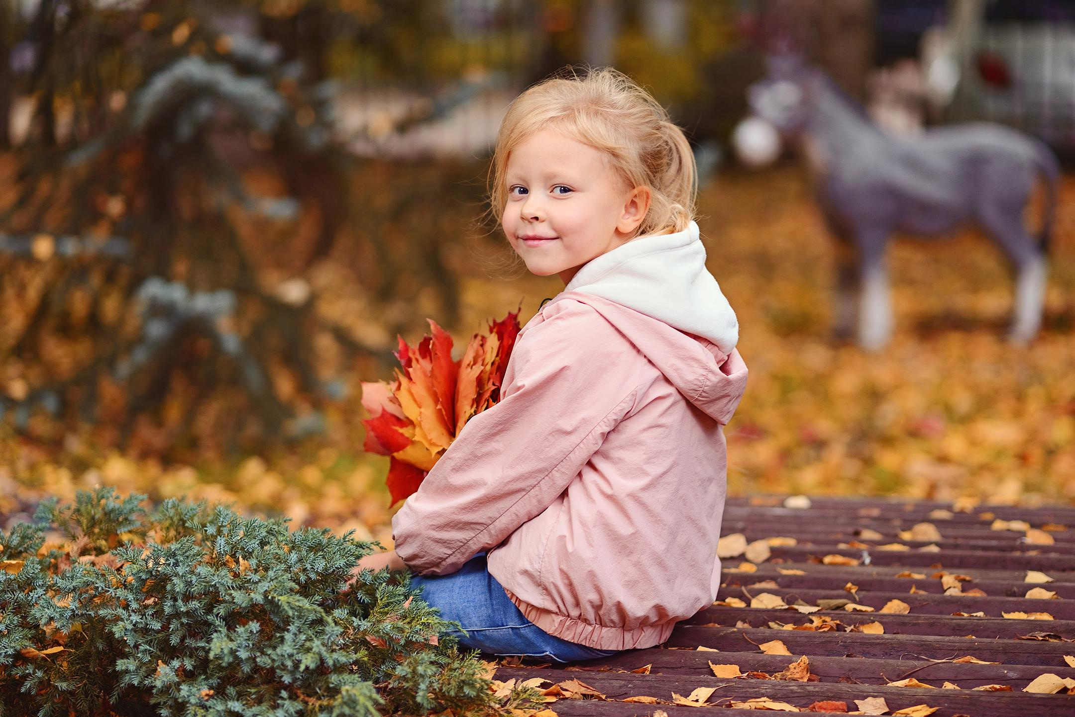 Портрет девочки с осенним букетом, сидящей на деревянном мостике во время прогулки в детском саду.