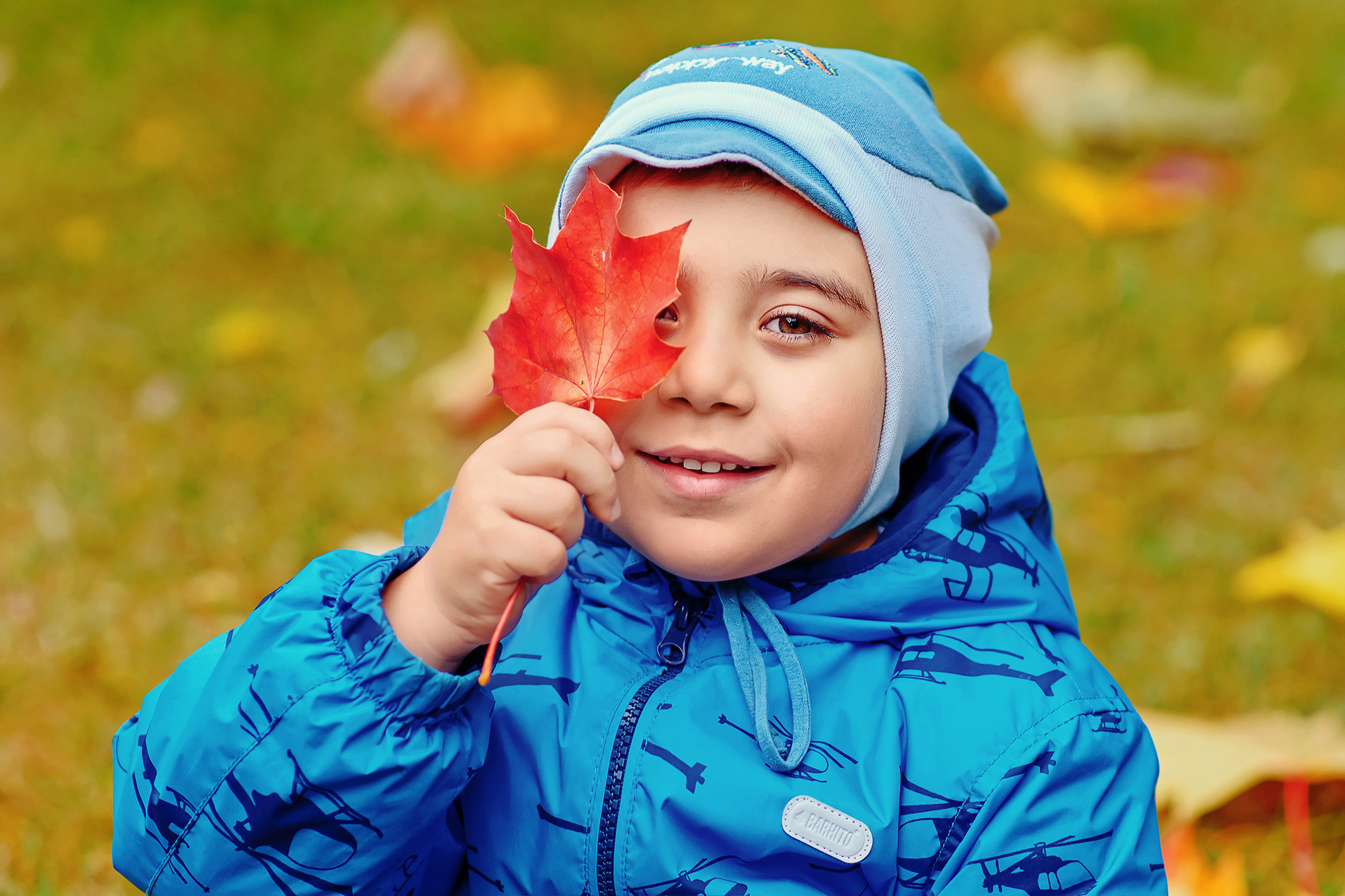 Мальчик на осенней прогулке в детском саду закрывает кленовым листиком один глаз.