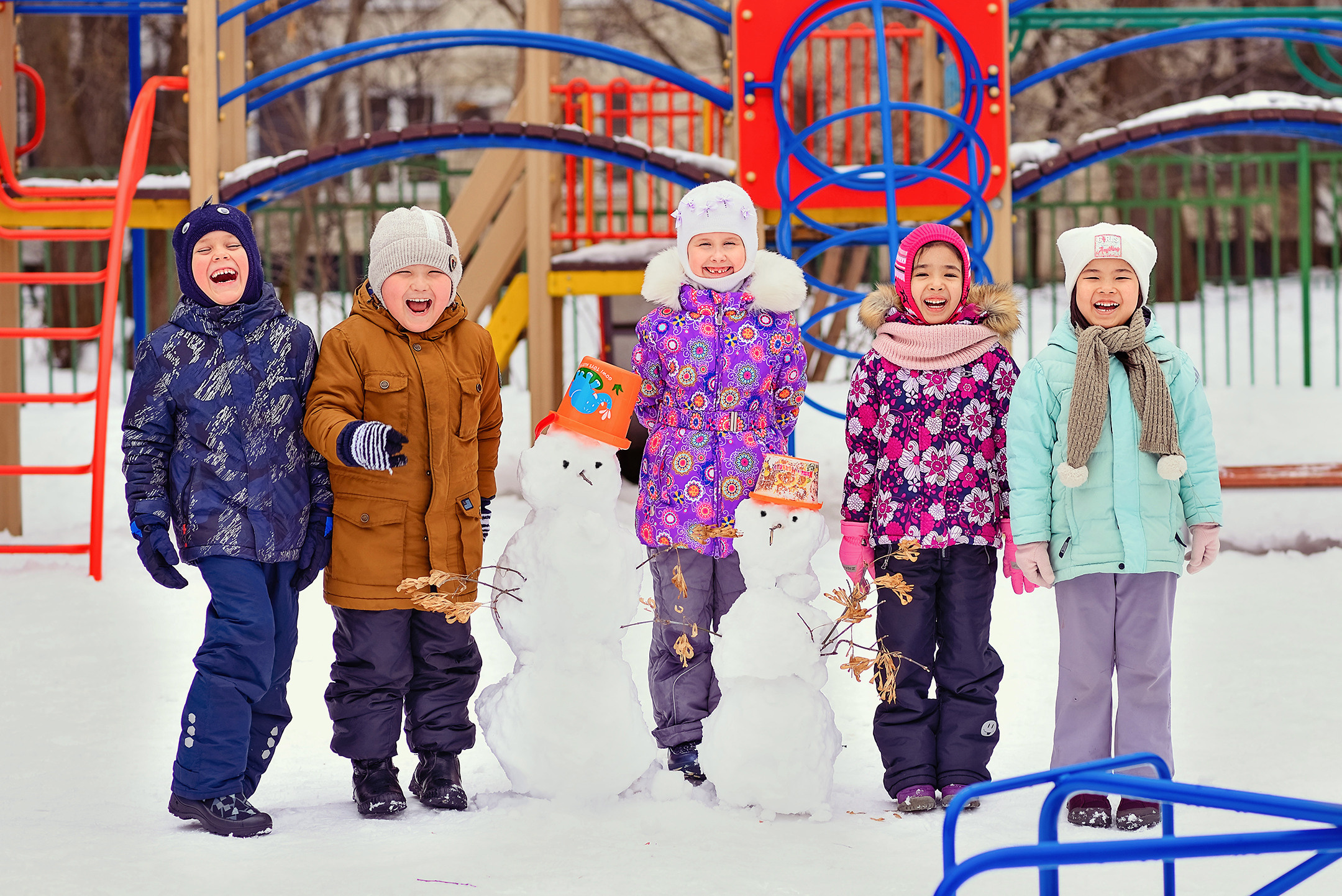 Пятеро смеющихся детей с двумя снеговиками на зимней прогулке в детском саду.