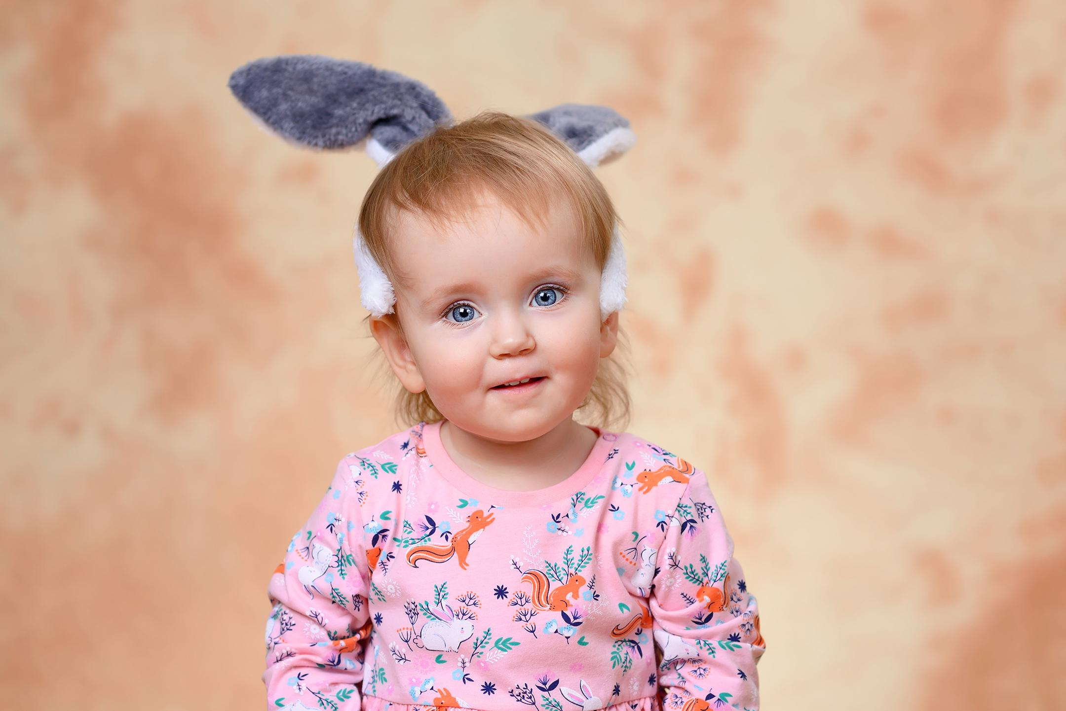 Игровой портрет улыбающейся девочки с заячьими ушками на голове на бежевом фоне в детском саду.