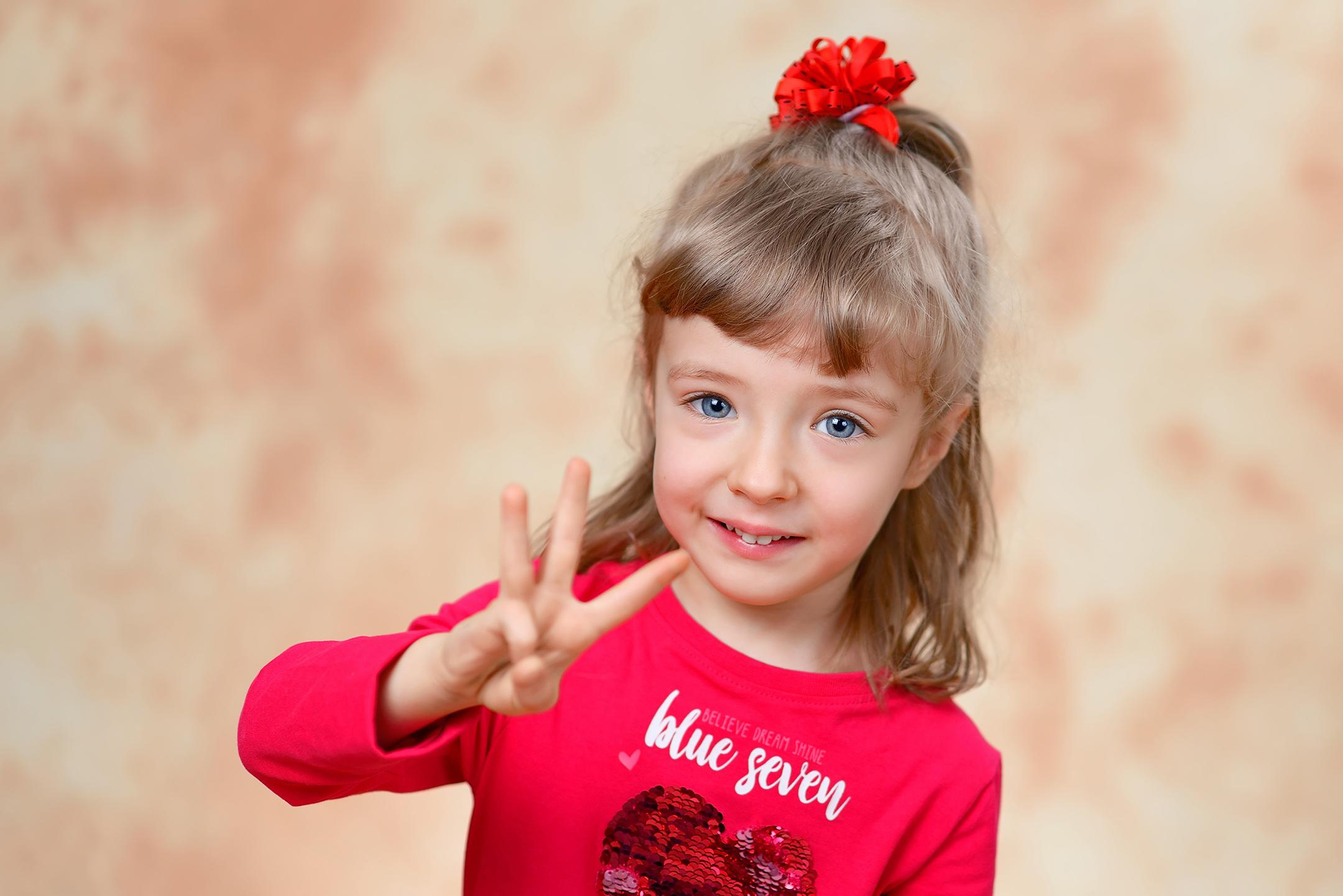 Портрет улыбающейся девочки в детском саду на студийном фоне, показывающей на пальцах свой возраст 3 года.