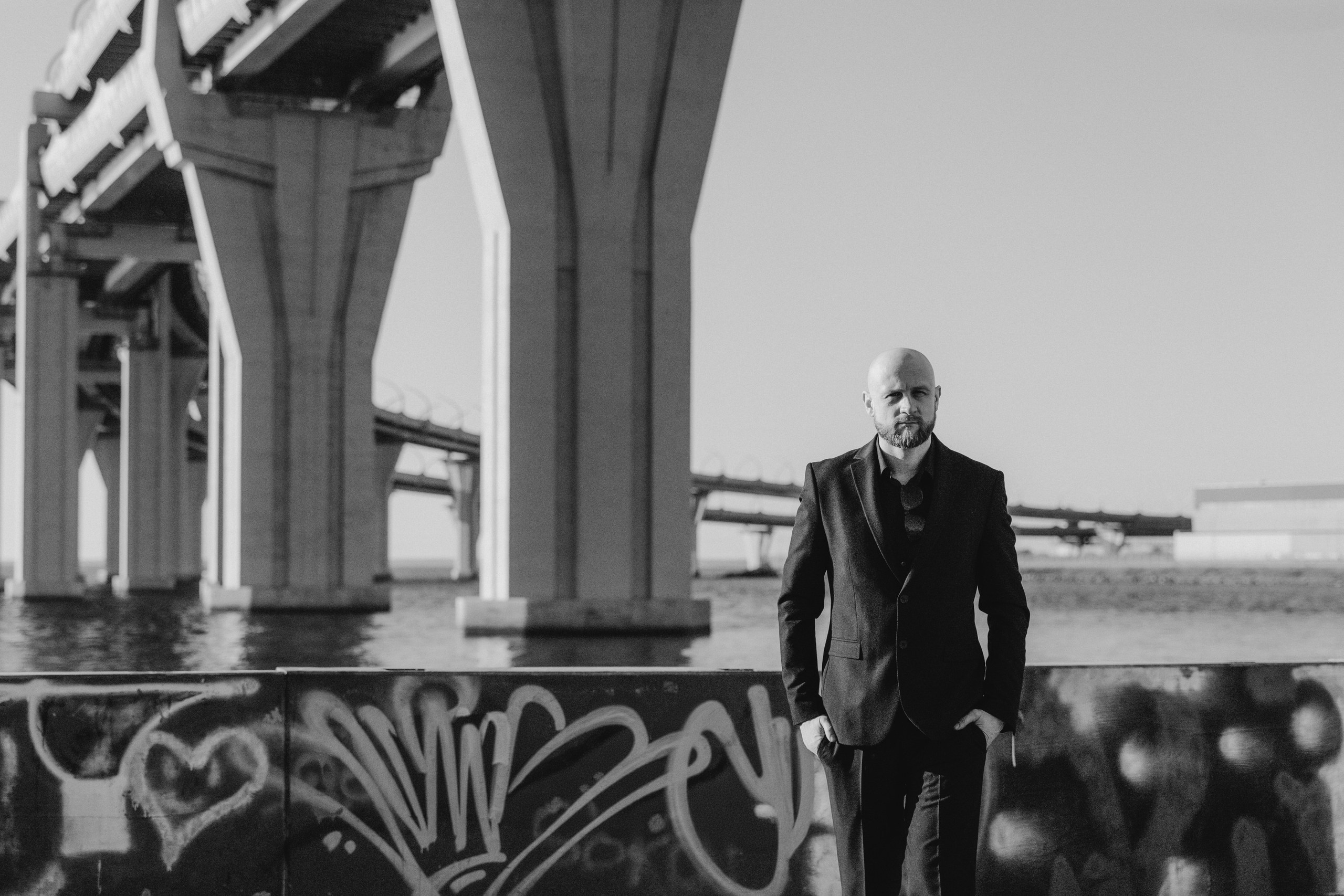 Мужчина в черном костюме позирует на фоне моста и граффити в Санкт-Петербурге, современный мужской портрет.