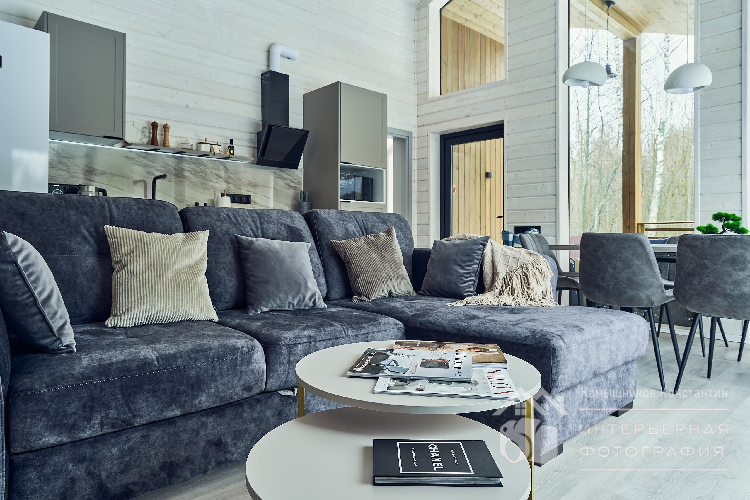 Интерьерная фотография гостиной в загородном доме: диван, журнальный столик, кухонная зона, панорамное окно, обеденный стол