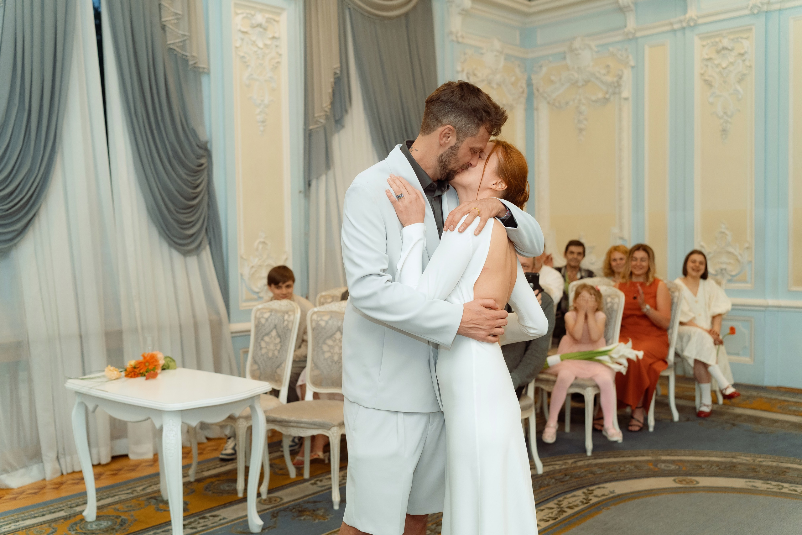 Первый поцелуй молодожёнов в голубом зале ЗАГСа, гости аплодируют позади