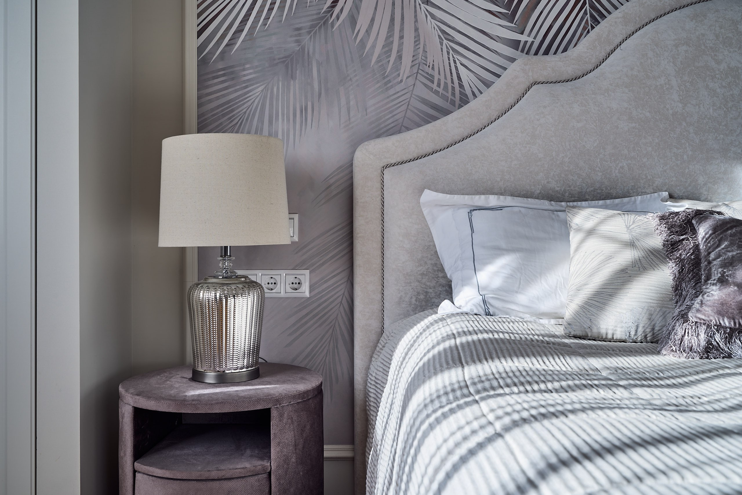 Интерьерная фотография мастер-спальни с прикроватной тумбой и частью кровати с подушками в солнечных лучах