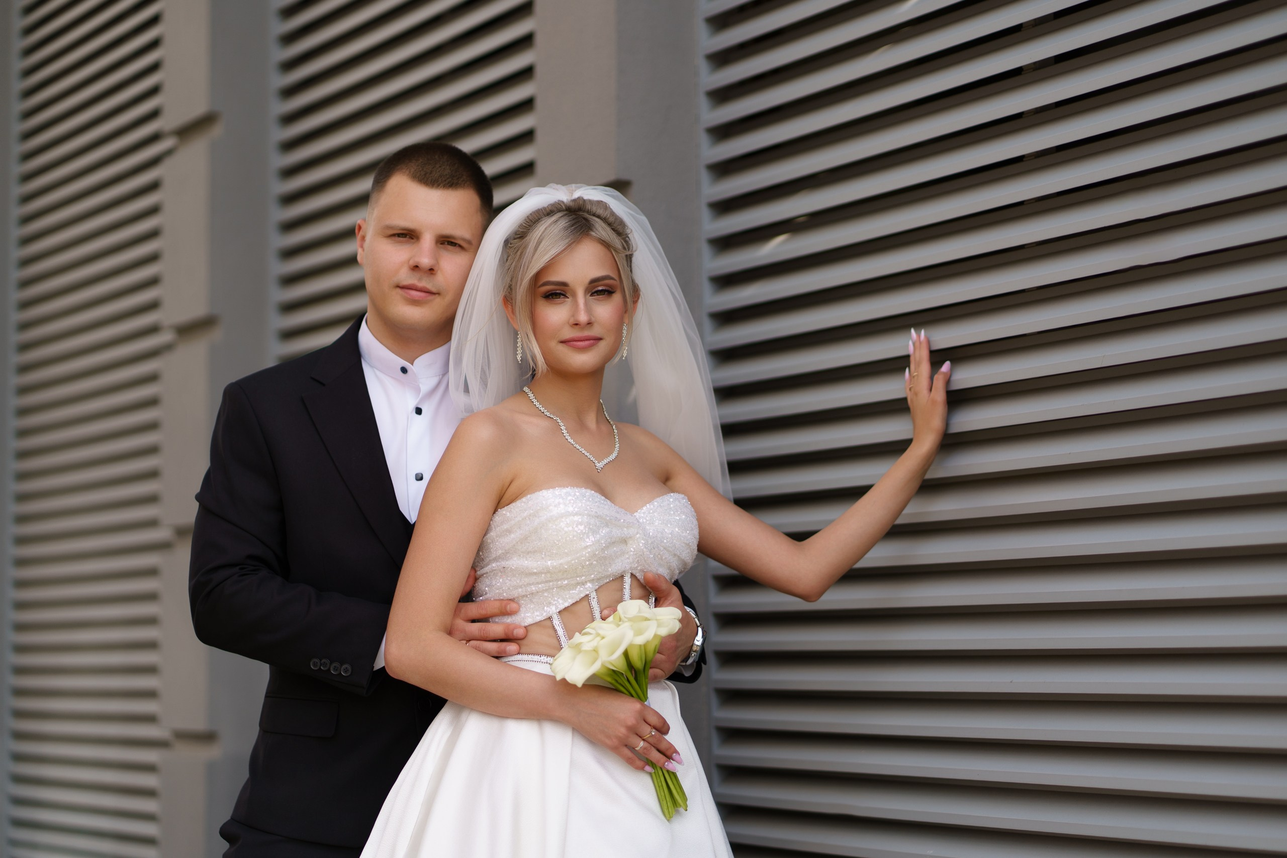 Архитектурный фон с рифлёными панелями подчёркивает красоту свадебной пары: невеста в белом платье с блестящими деталями, жених — в классическом чёрном костюме | свадебная фотография для альбома Екатеринбург