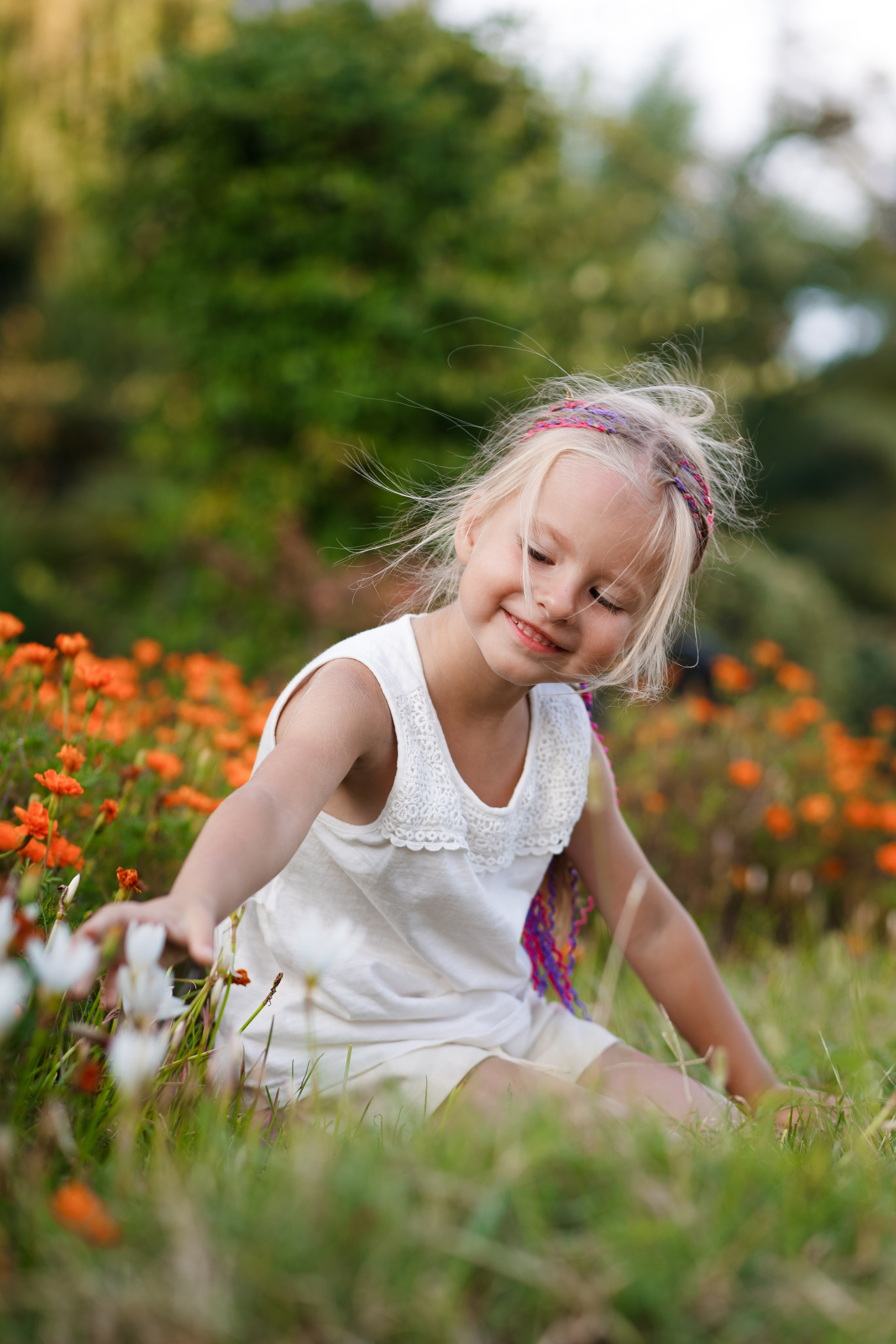 Маленькая девочка с белыми волосами сидит на клумбе с цветами и улыбается