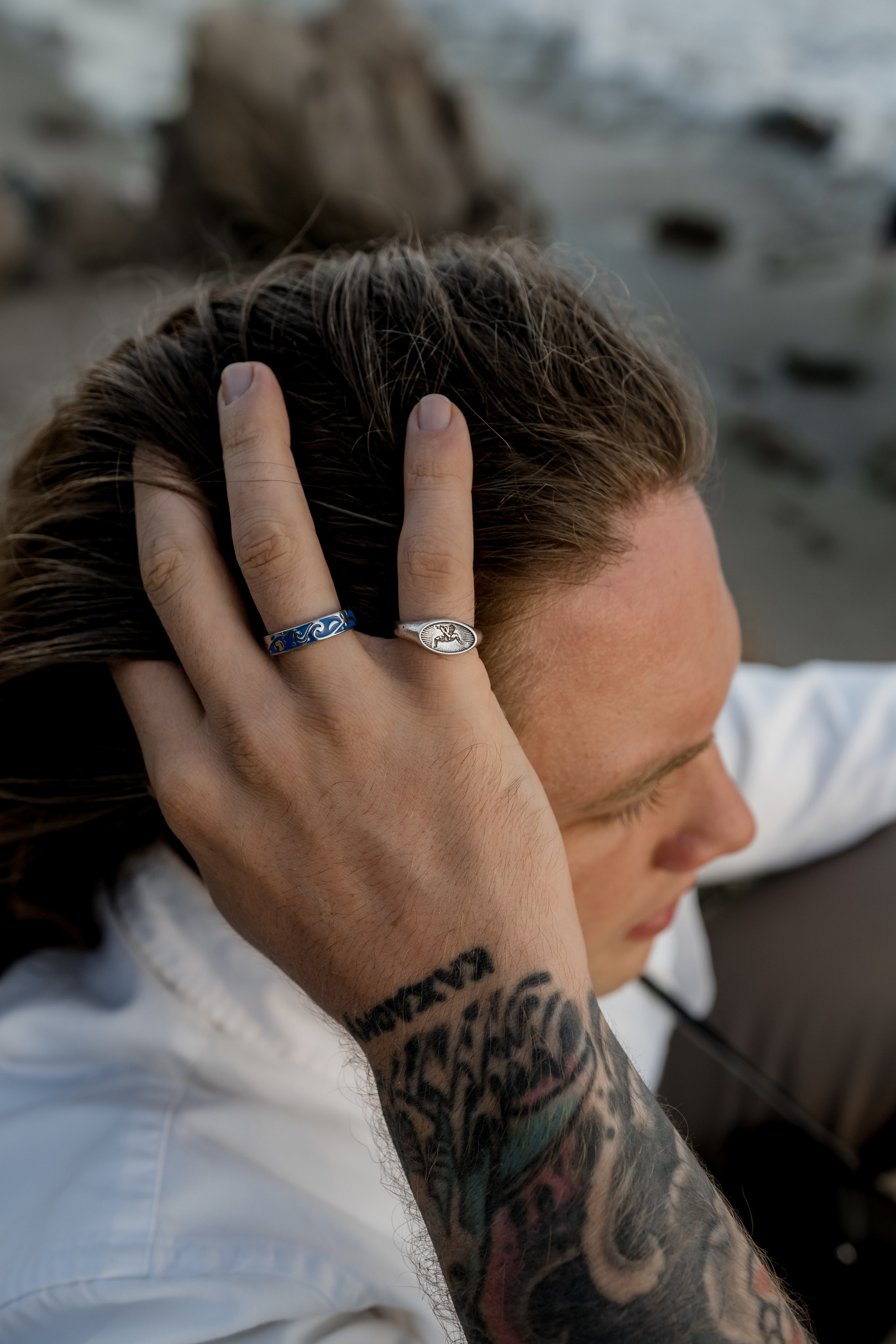 Male model with tattoos styled in casual wear, running his hand through his hair by the seaside.