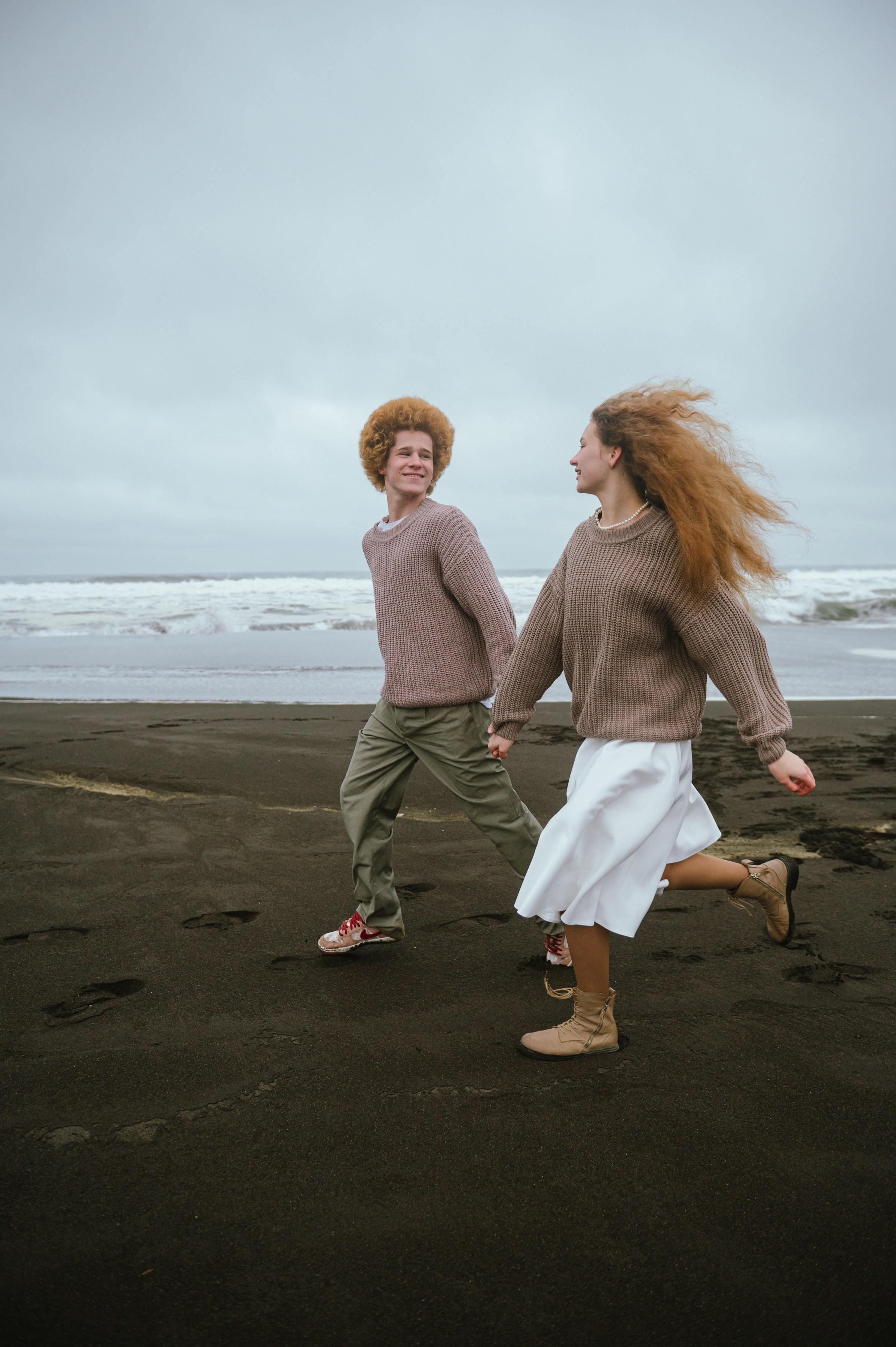 Парень и девушка бегут по чёрному пляжу у моря, волосы развеваются на ветру. Атмосфера свободная, весёлая.
