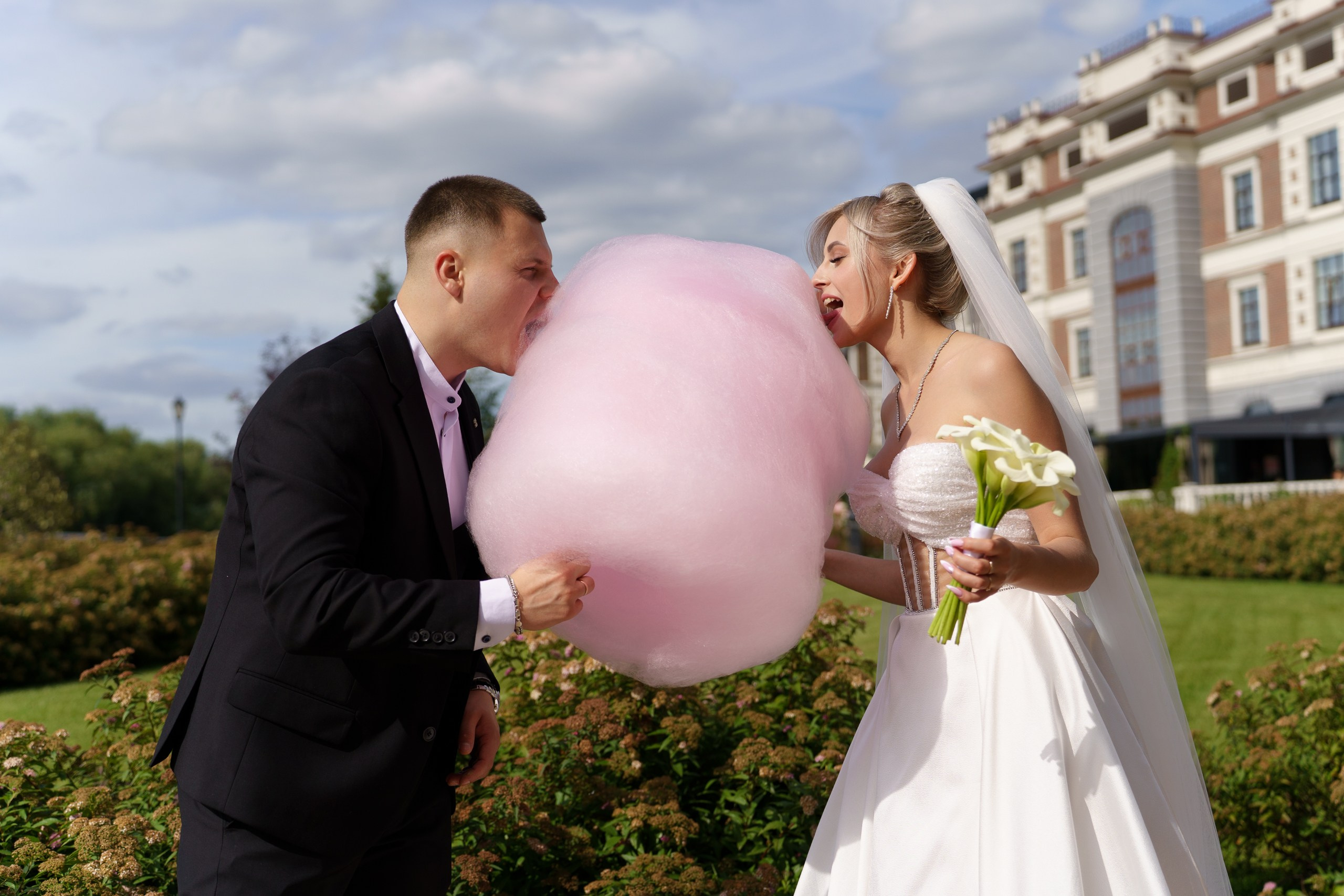 Весёлый момент со свадьбы: жених и невеста с улыбками пробуют огромную сахарную вату. Невеста держит букет, её наряд дополнен фатой, жених выглядит элегантно в классическом костюме | фотограф для креативной свадьбы Екатеринбург