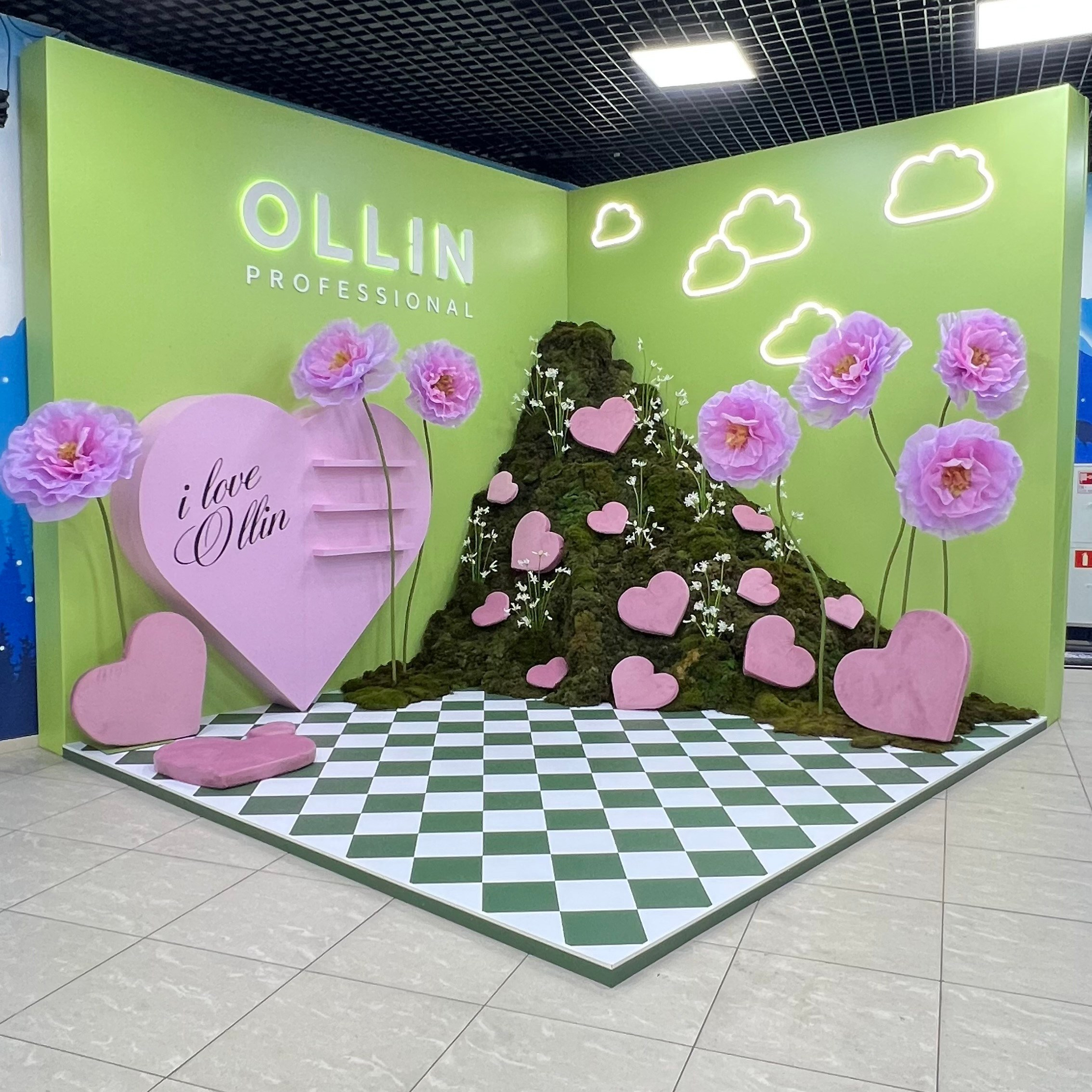 Угловая фотозона бренда «OLLIN PROFESSIONAL» в природно-весеннем стиле. Стены конструкции окрашены в ярко-зеленый цвет и дополнены светящимися неоновыми контурами облаков. На полу уложено шахматное зелено-белое покрытие. Композиция включает холм из зеленого мха, усыпанный розовыми сердечками разного размера, и гигантские декоративные цветы магнолии на длинных стеблях. Слева установлено большое сердце с надписью «i love Ollin».