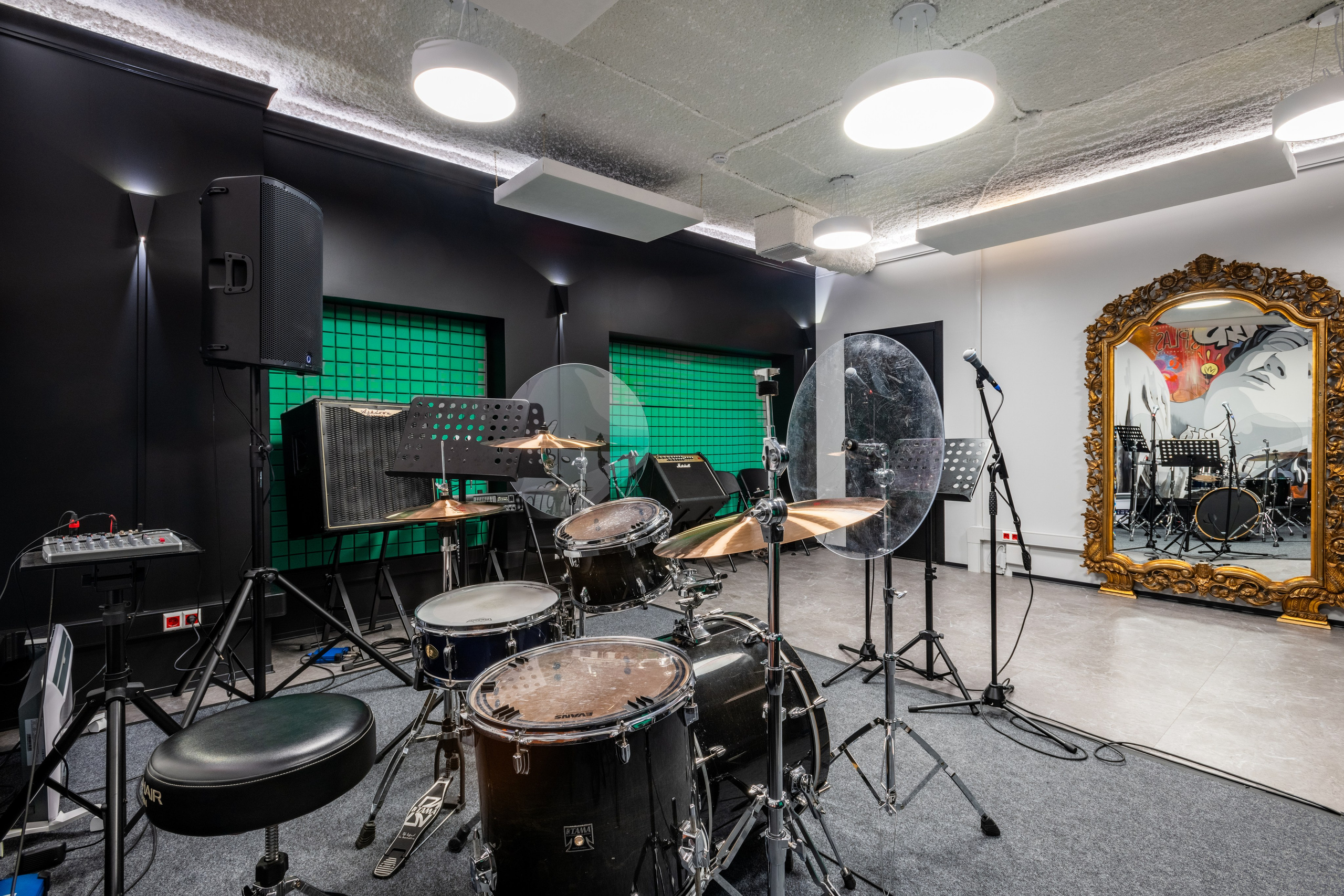 Пространство для репетиций с зеленой светодиодной подсветкой, набором ударных, гитарами и большим зеркалом в раме на стене.