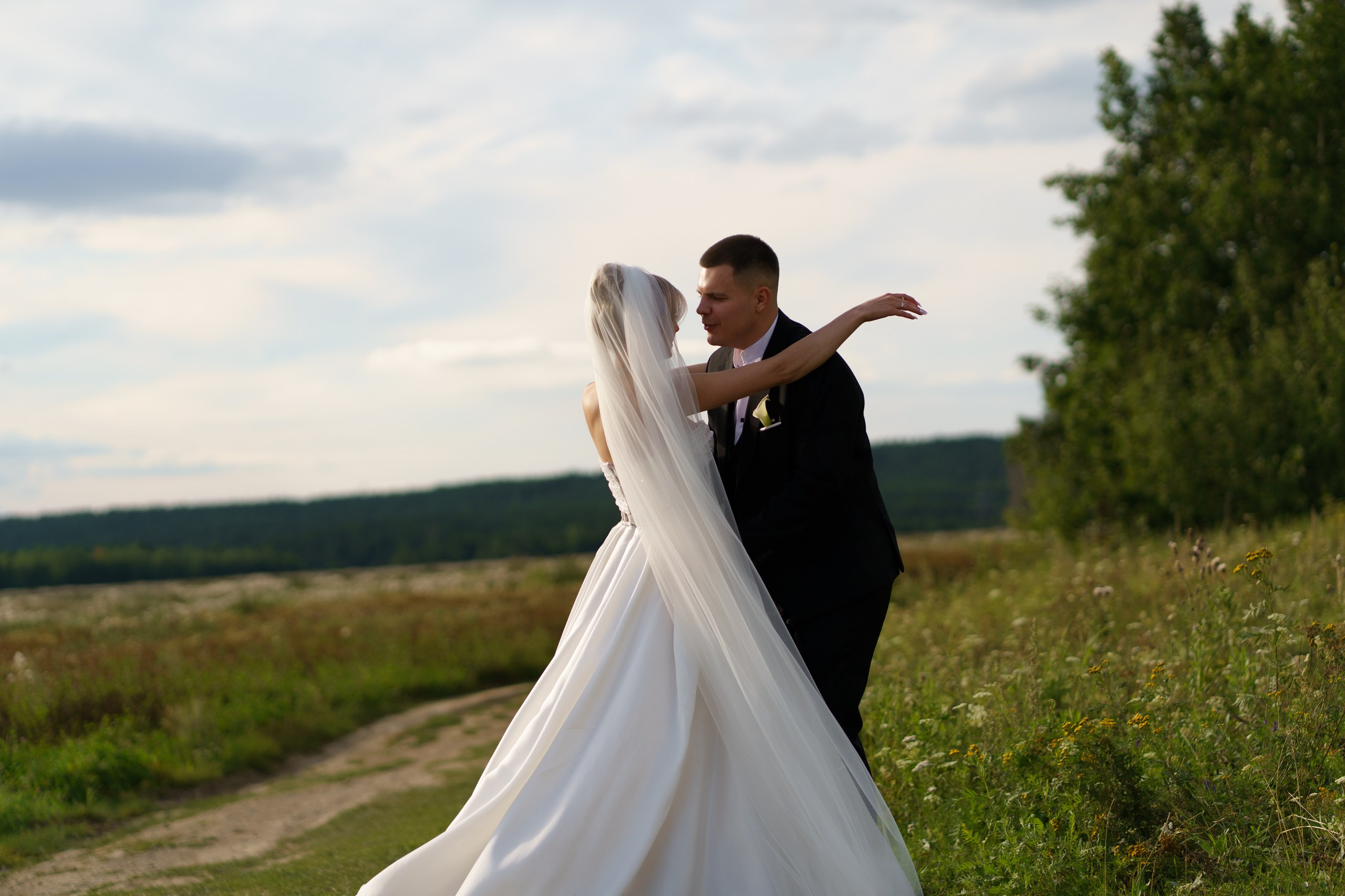 Невеста в роскошном белом платье с длинной фатой и жених в чёрном костюме с бутоньеркой стоят на просёлочной дороге. Вокруг — высокая трава, полевые цветы и зелёные деревья, на небе лёгкие облака | фотограф для красивых свадеб Екатеринбург