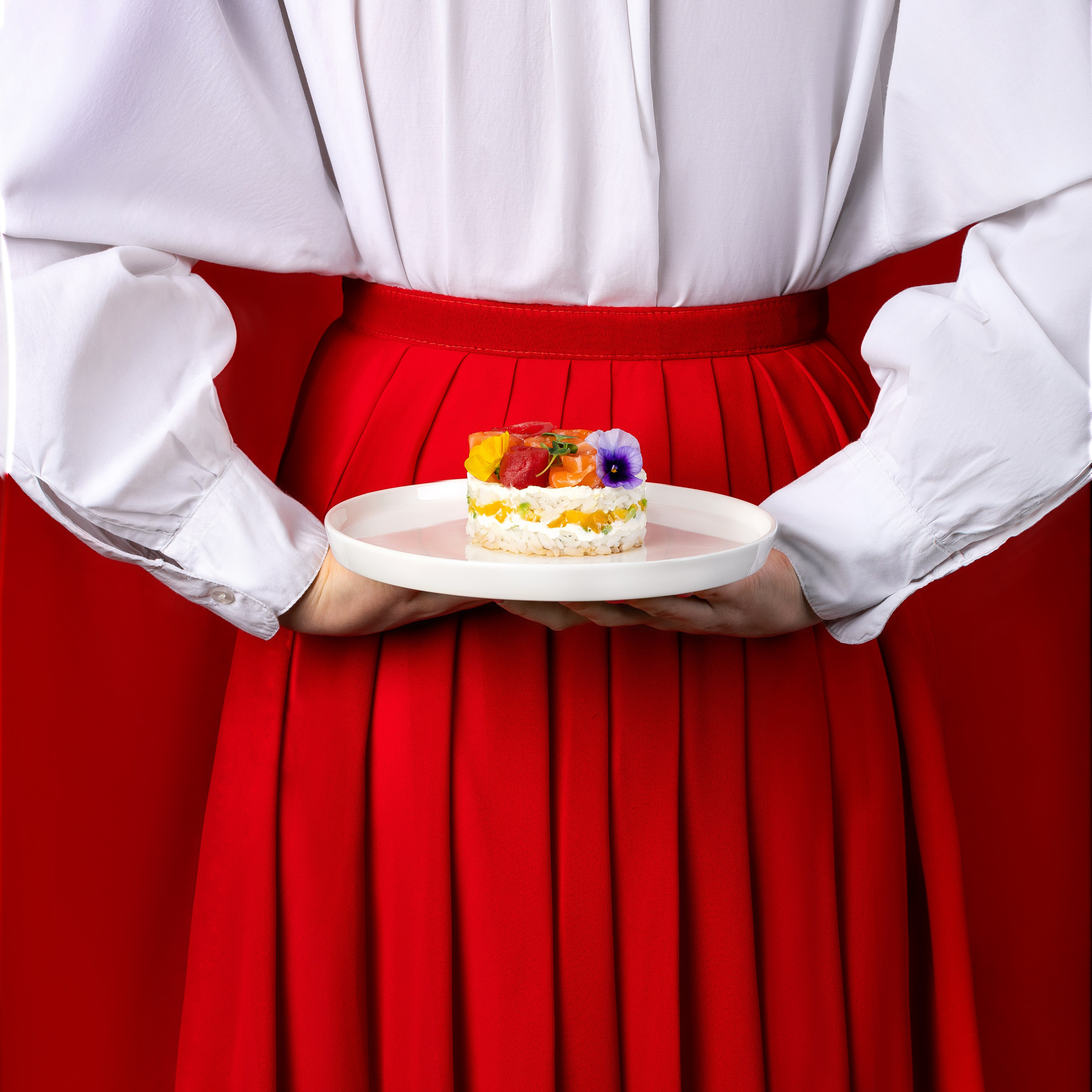 Студийная съёмка для ресторана 'ТАНУКИ': женщина в красной юбке и белой рубашке держит тарелку с десертом, украшенным съедобными цветами. Фото сделано на фоне красного фона, подчеркивающее яркие и свежие цвета блюда.