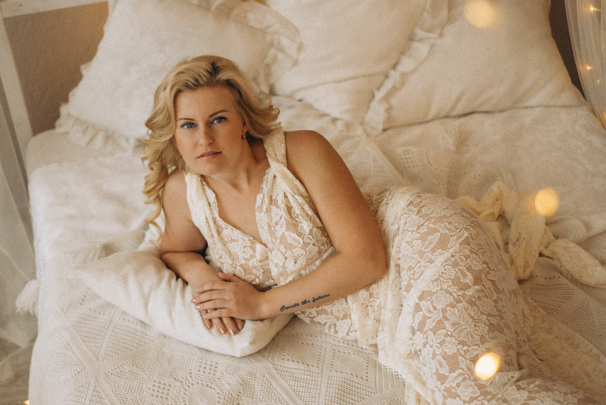 Натали Каролин лежит на животе на белой кровати. Вид сверху, модель в кружевном платье, татуировка "Create the future" на предплечье. Атмосферная фотосессия в стиле бохо. Фотограф Василий Дан