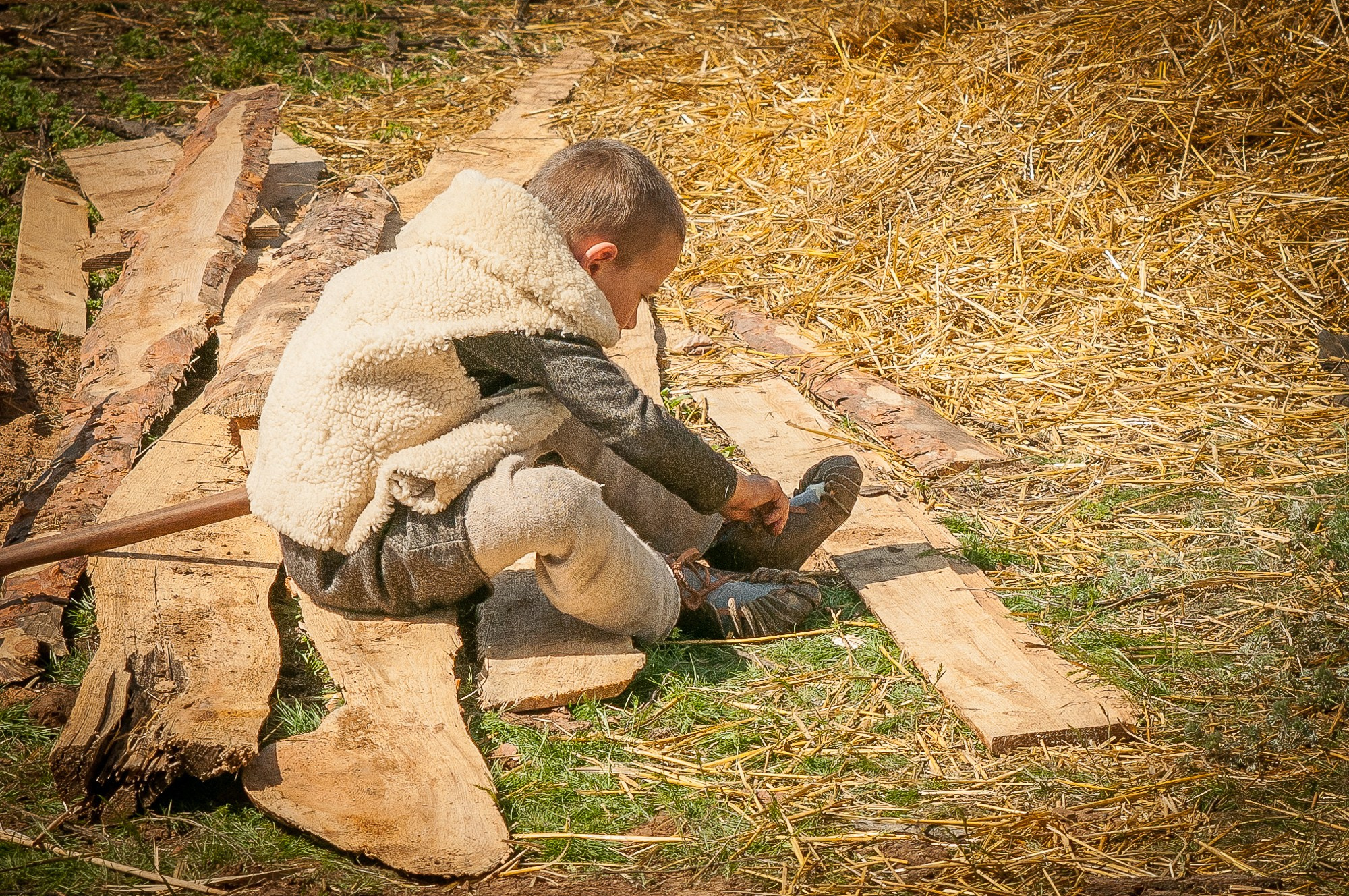 Мальчик в жилетке из белой овчины надевает старинные ботинки сидя на земле